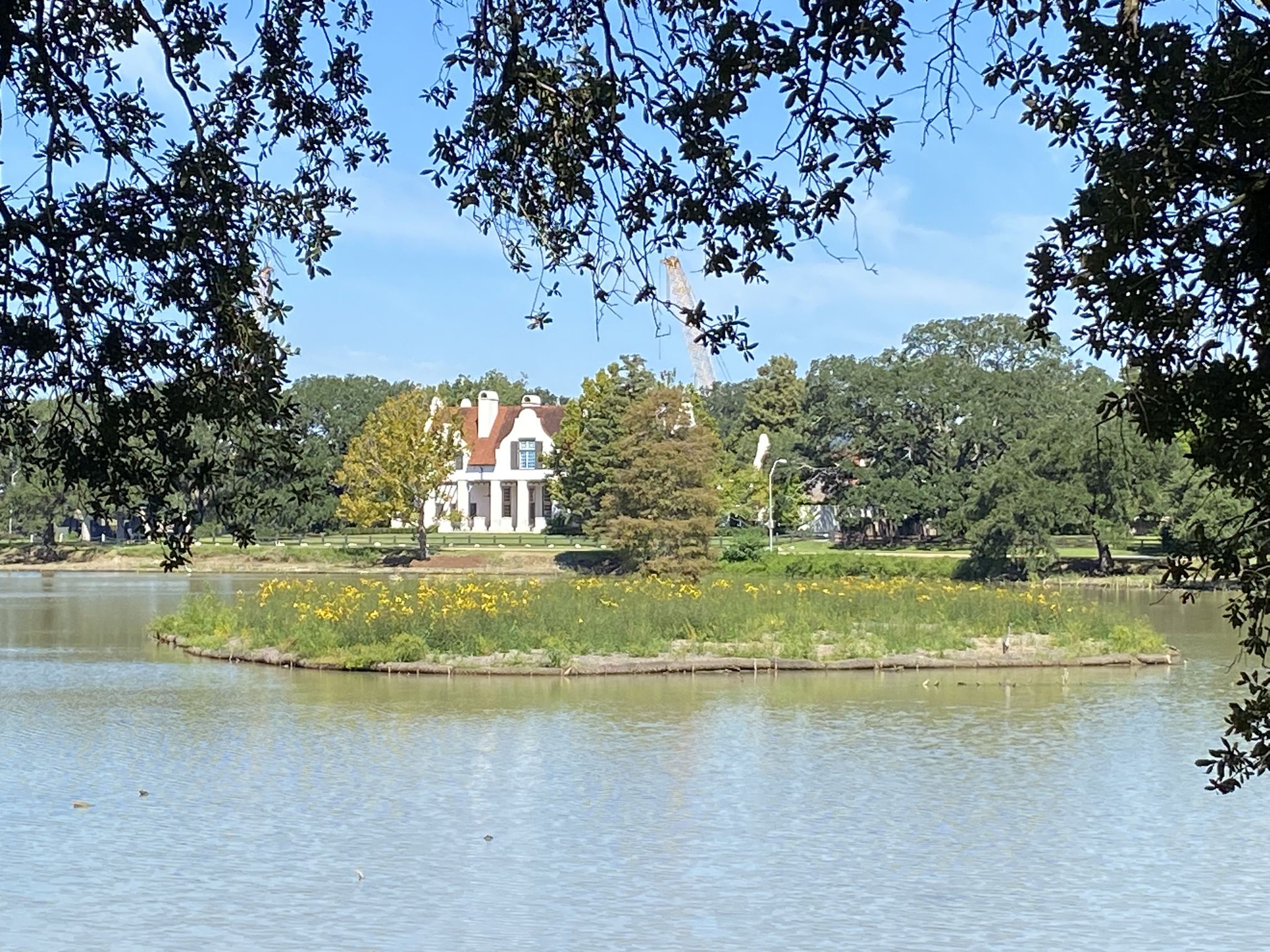 A large house on a small island in a lake, framed by tree branches. - City-Brooks Community Park – City Park Lake loop from LSU Rural Life Museum