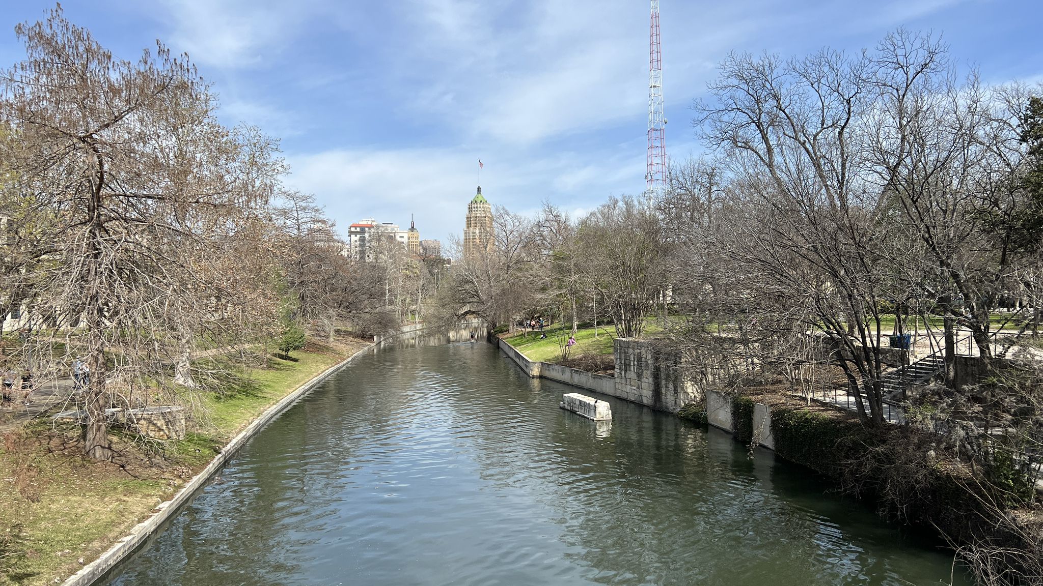 A calm river flows through a park with bare trees and distant buildings. - Bexar County Courthouse – San Antonio River Walk loop from San Antonio