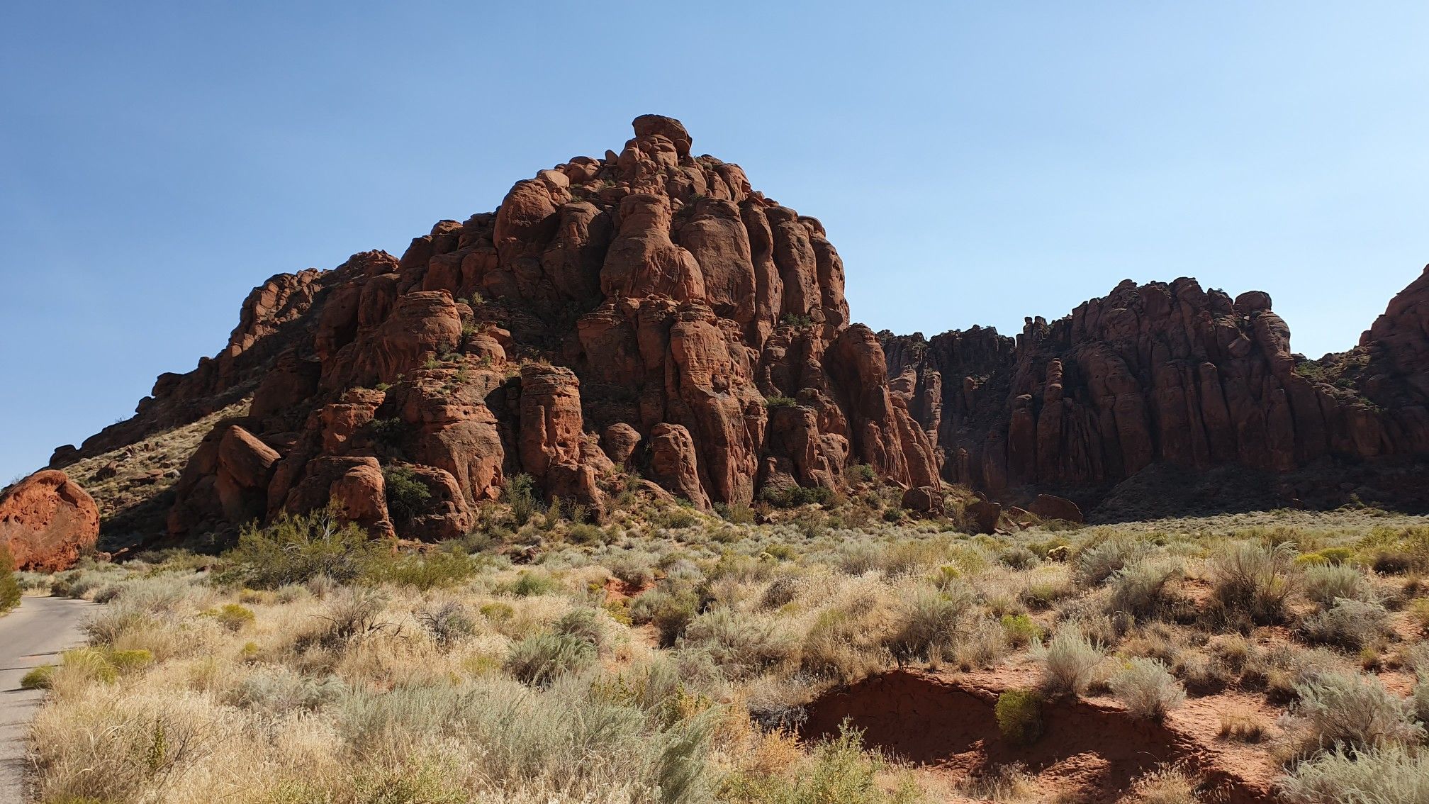 Massive red rock formations rise above desert shrubs and a paved road. - Snow Canyon loop