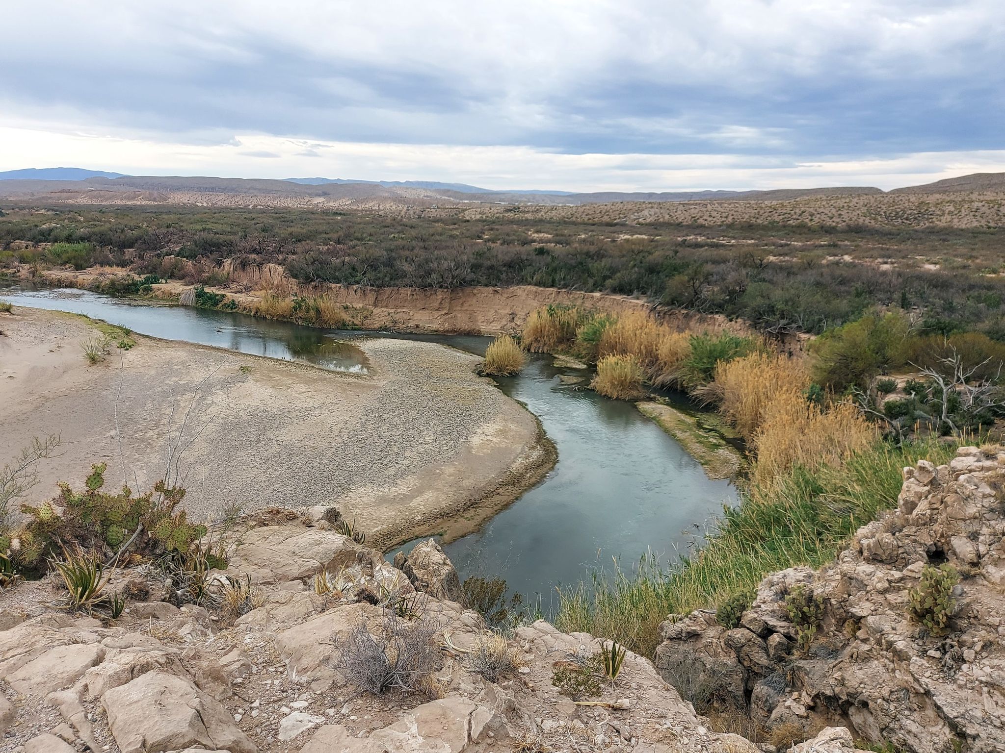 A river with sandy banks and green reeds flows through an arid landscape. - Boquillas Canyon Trail