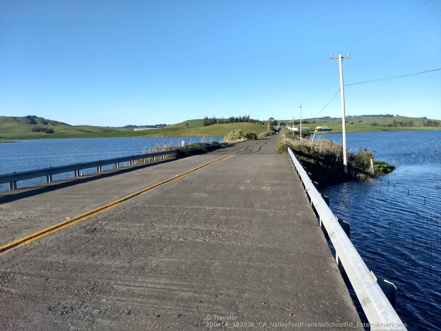 A sunny day shows a road crossing calm blue water with distant green hills. - Estero Americano loop from Valley Ford