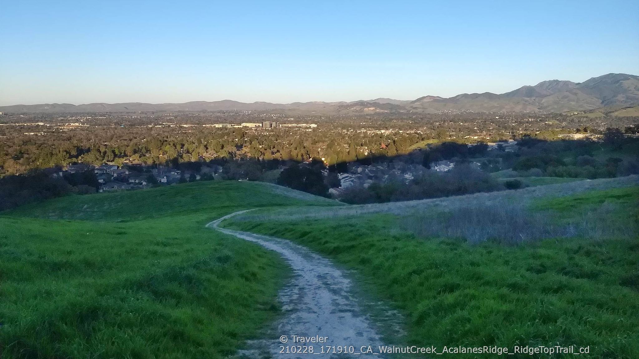 A winding dirt path through lush green hills, with a distant city and mountains. - Camino Verde and Ridge Top Trail Loop