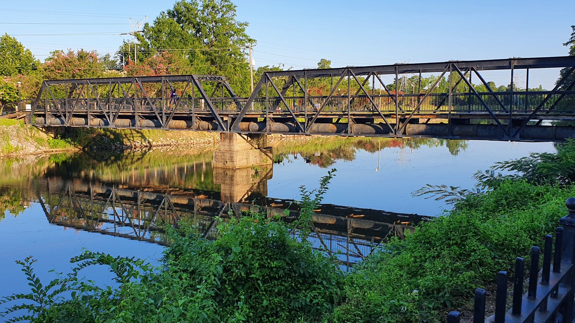 Metal truss bridge over a calm river, reflecting trees and sky, in warm light. - Riverfront Park Canal Greenway