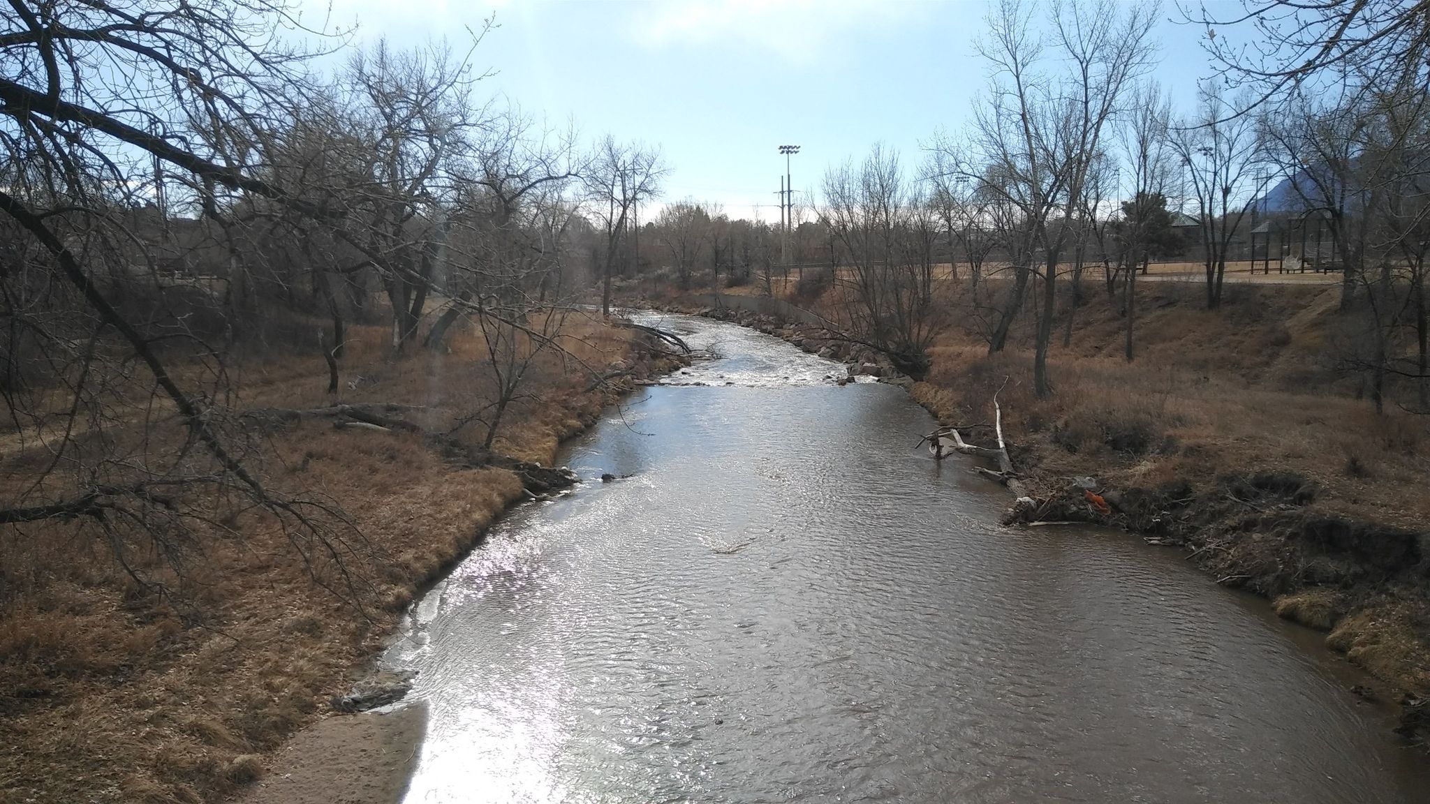 A river flows through a dry, treed landscape under a bright sky. - Uintah street bridge – Pikes Peak Greenway loop from Colorado Springs