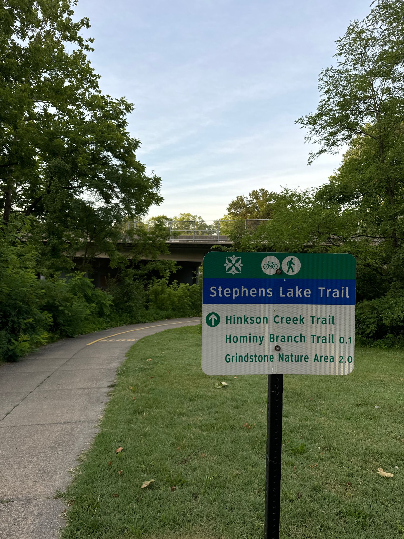 A trail sign stands next to a paved path surrounded by green trees. - Stephens Lake loop from Columbia