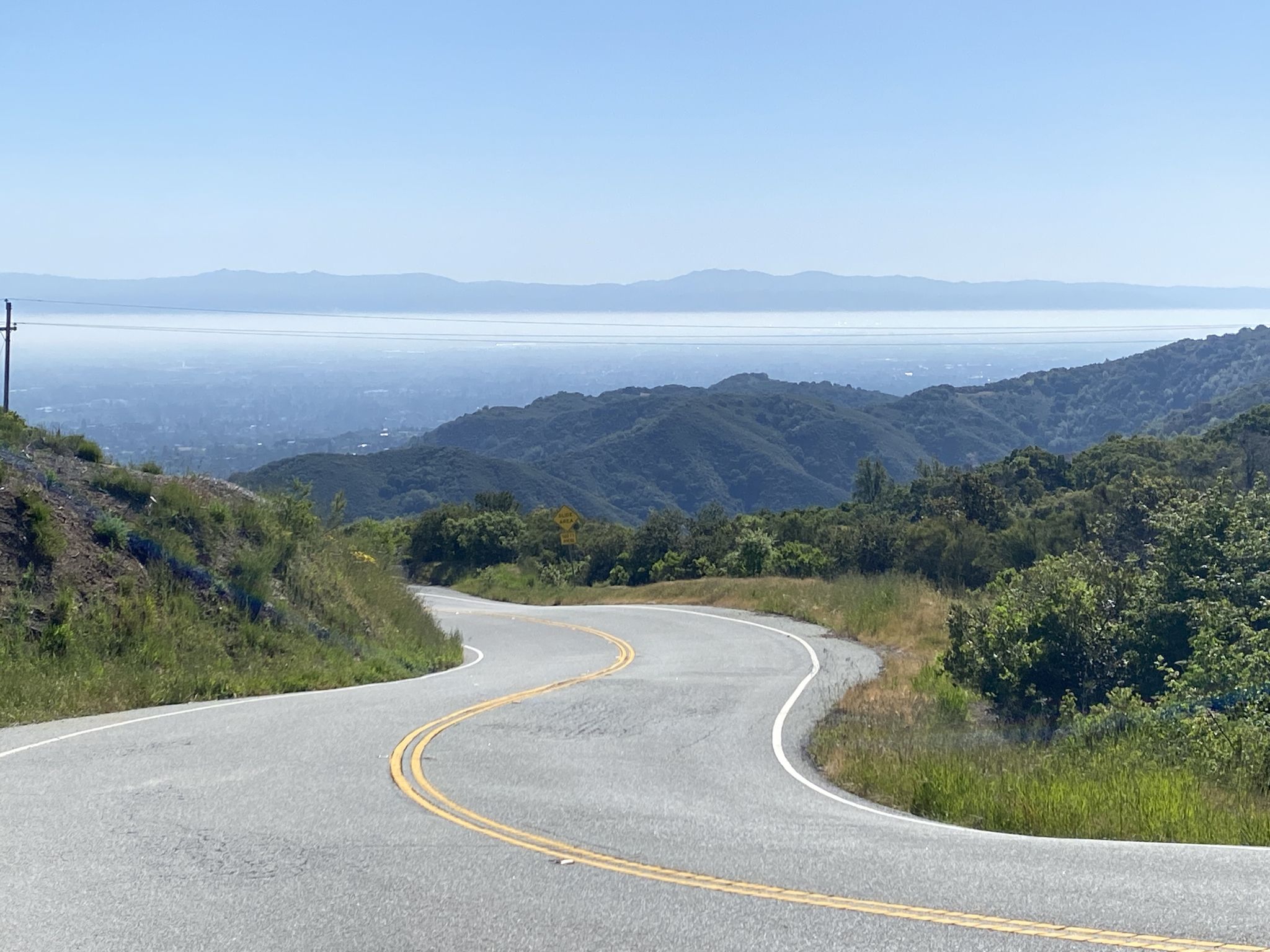 A winding road descends through green hills, overlooking a distant hazy valley. - La Honda & Skyline Ridge loop from Mountain View