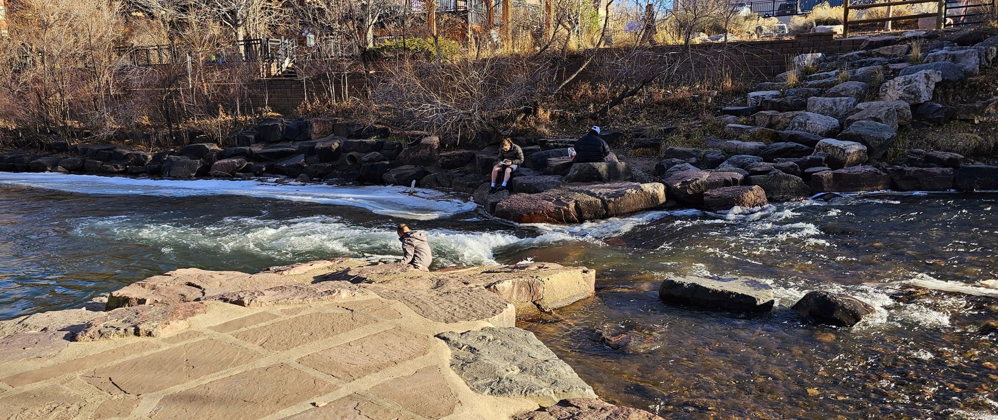 Two people sitting on rocks by a rushing river with distant buildings. - Clear Creek Trail – Fairmont Canal Trail loop from Arvada West Dog Park