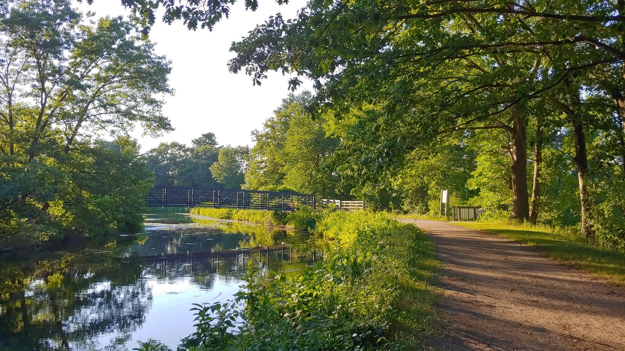 Sunny canal with a dirt path alongside, surrounded by lush green trees. - Beginner Loop