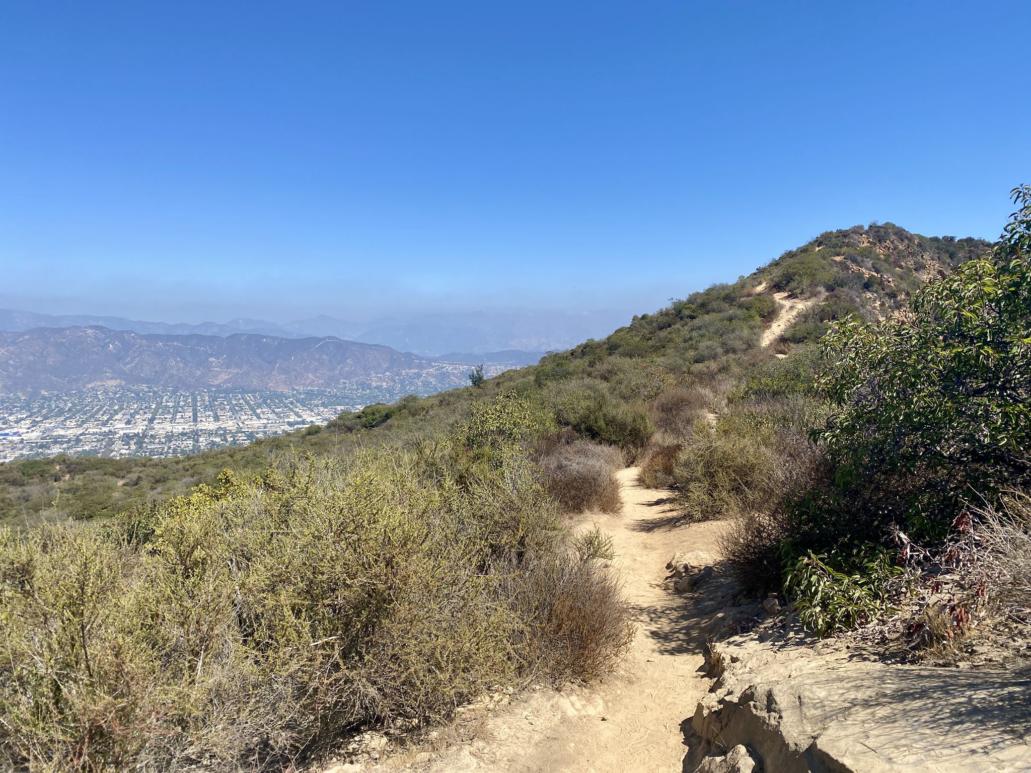 Dirt trail winding up a brush-covered hillside with a city in distance. - Burbank Peak, Cahuenga Peak and Mount Lee Loop