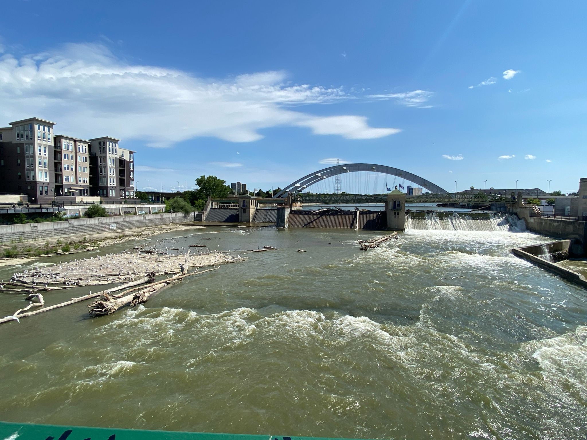 Sunny urban river scene with a dam, arched bridge, and distant buildings. - Genesee Riverway Trail