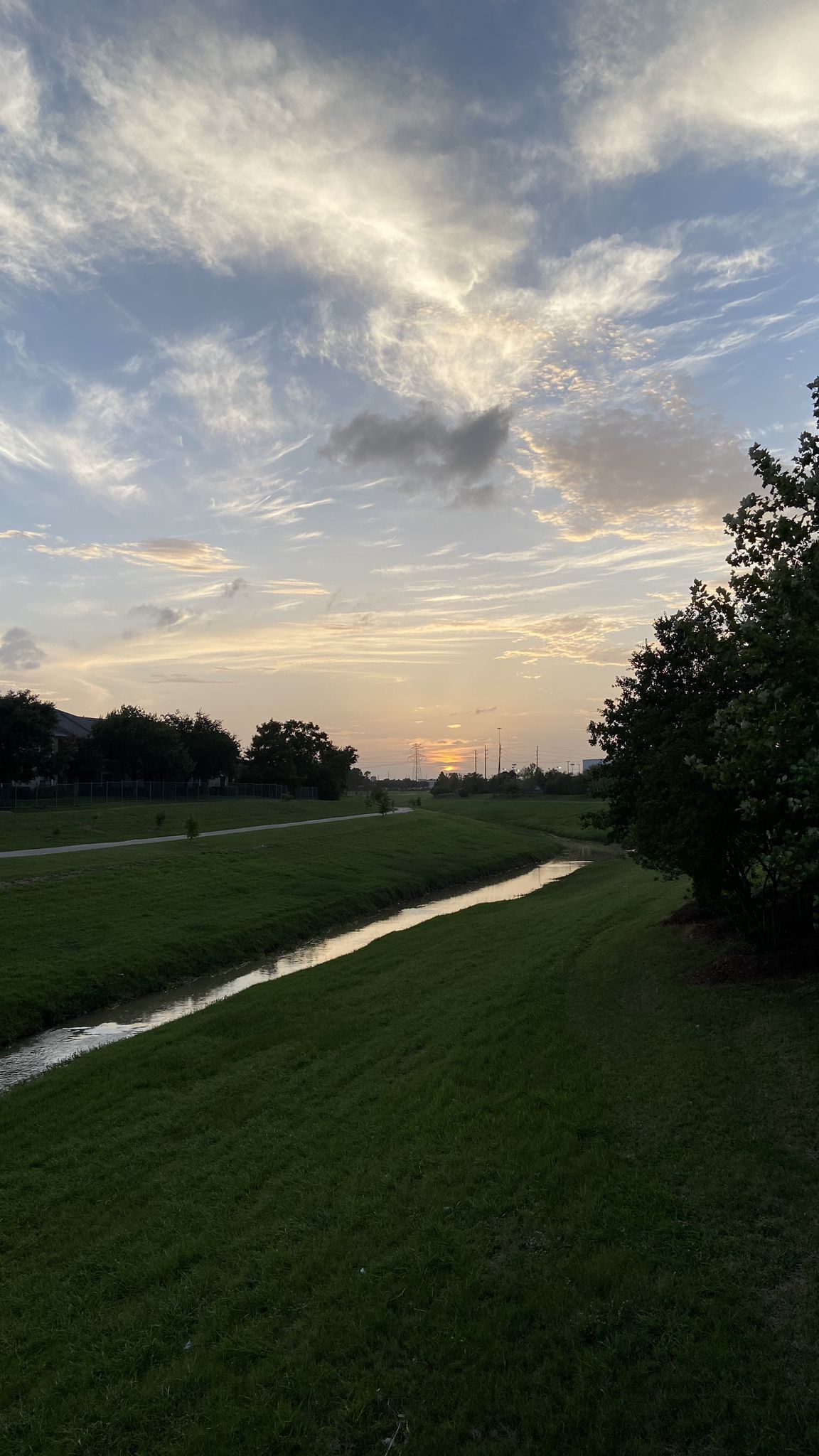 A narrow stream flows through green fields under a cloudy sunset sky. - Rick Rice Park loop from James E Taylor High School