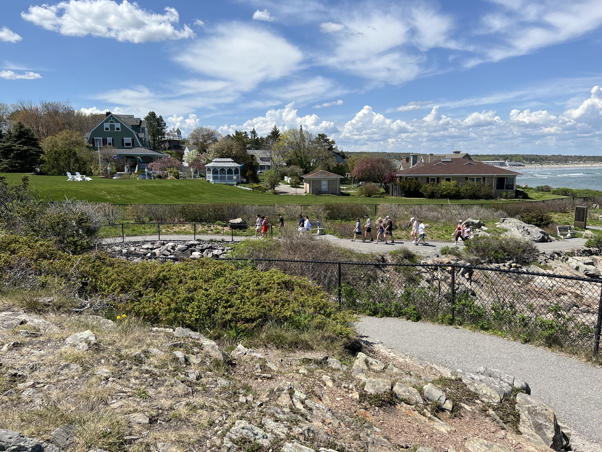 People walk on a path along a grassy hill with houses and ocean. - Marginal Way