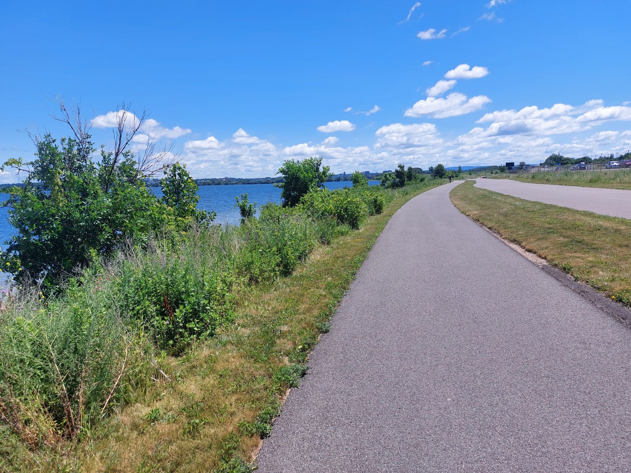 Paved path running alongside a large blue lake with green vegetation on a sunny day. - Onondaga Lake West Shore Trail