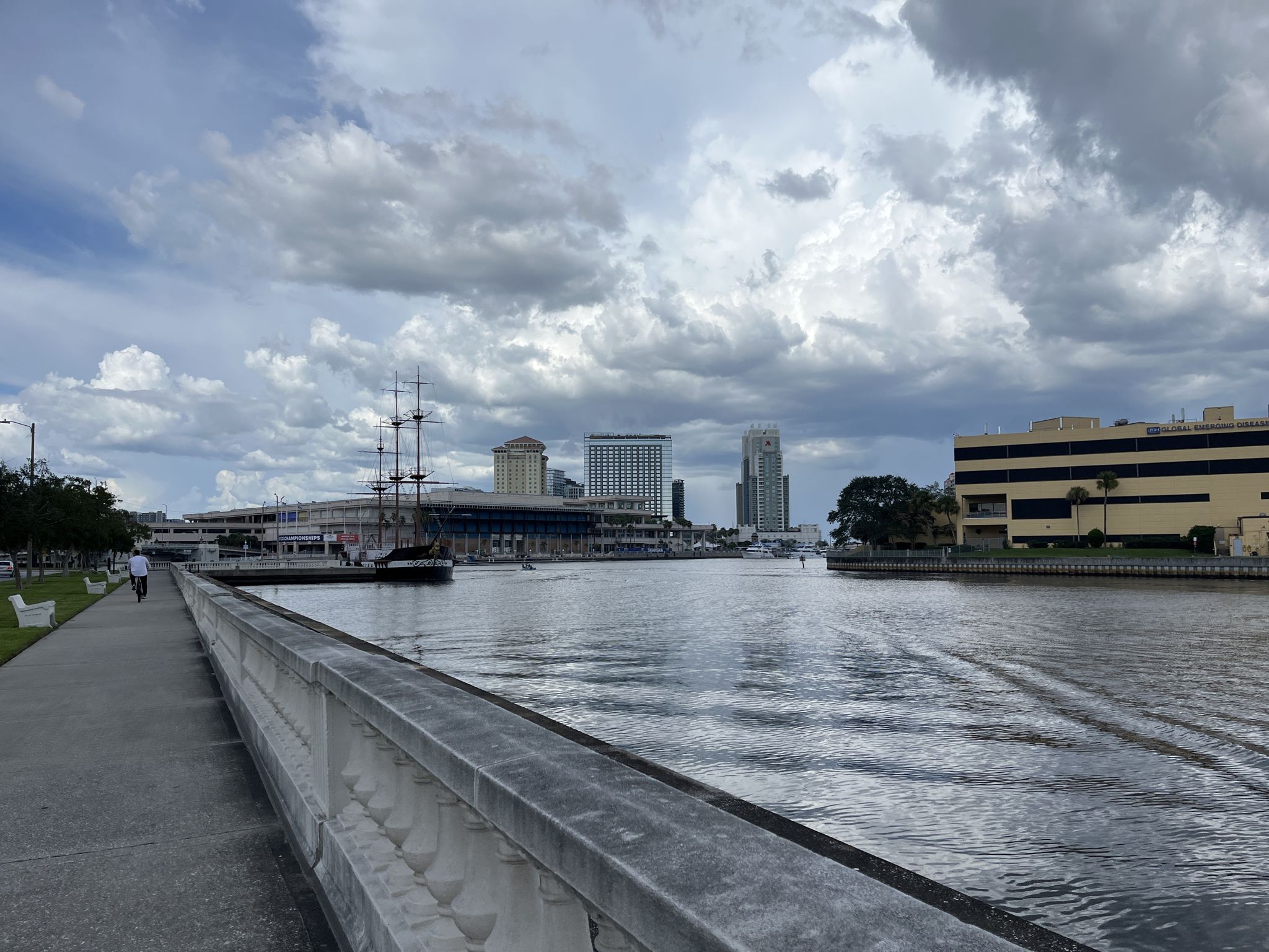 Riverfront walkway with city buildings and a sailboat under a cloudy sky. - Bayshore Boulevard