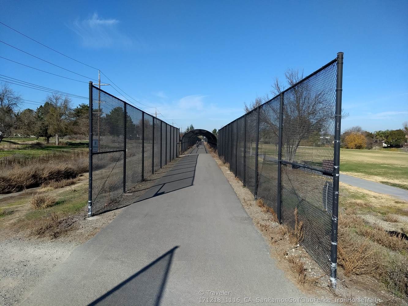 A paved path extends between two tall chain-link fences under a clear sky. - Golf Ball Protection Tunnel – Iron Horse Trail loop from Dublin High School