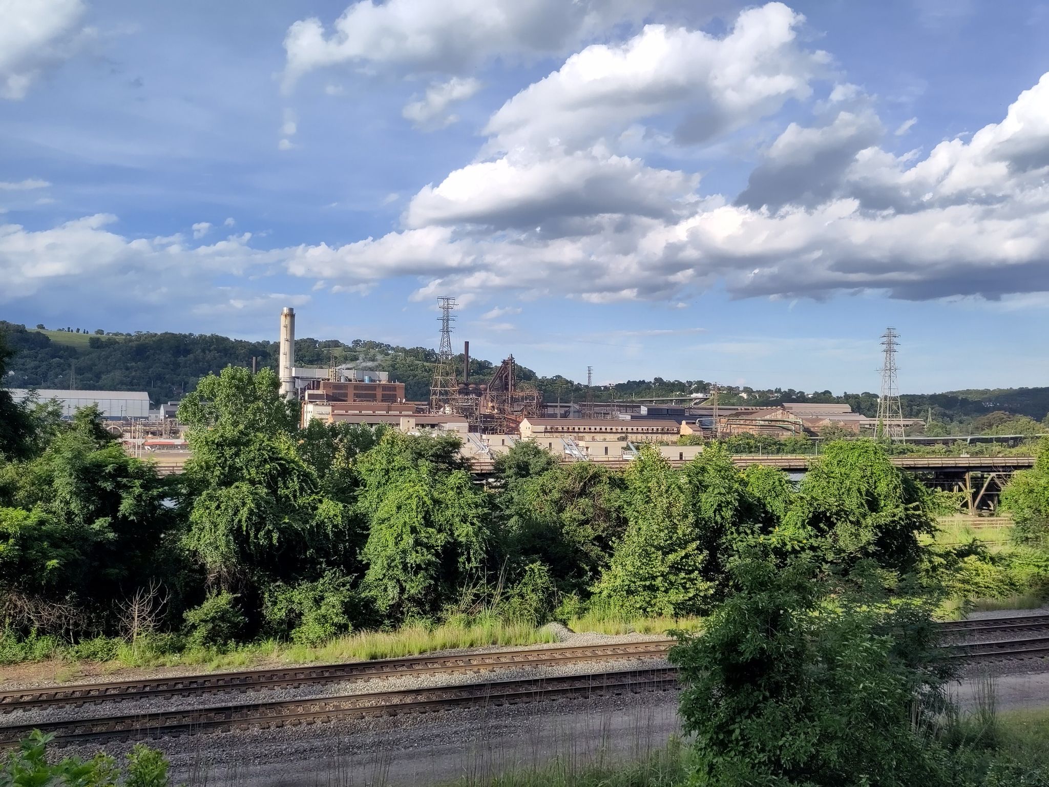 An industrial complex with smokestacks is visible behind train tracks and trees. - Steel Valley Trail