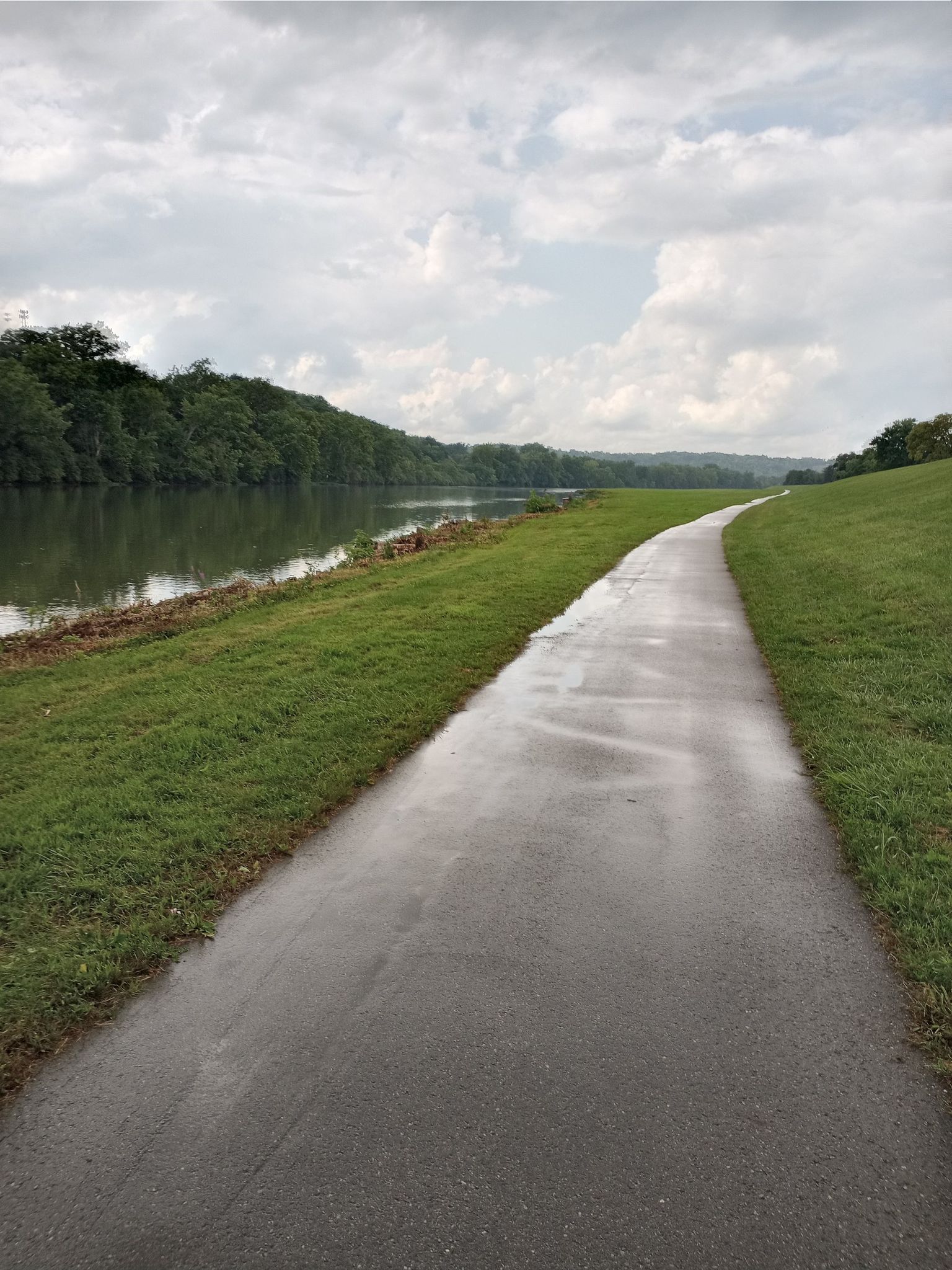 Wet paved path alongside a river with green grass under a cloudy sky. - Miamisburg Riverfront Park – Hutchins State Reservoir loop from West Carrollton