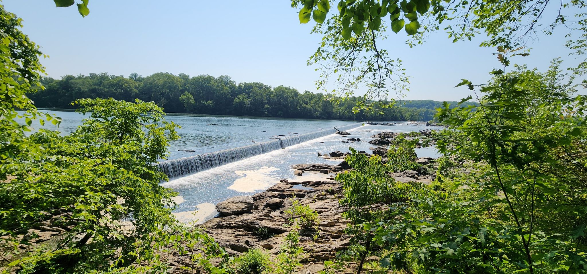 Sunny river view with a low dam creating a waterfall, framed by green trees. - Potomac Heritage National Scenic Trail
