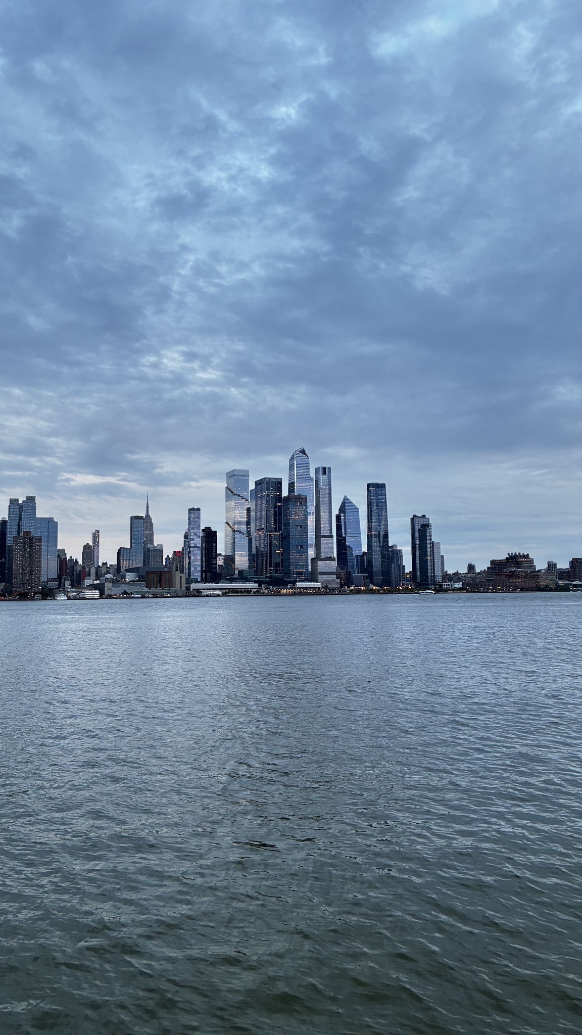 Modern city skyline across a wide river under a dramatic cloudy sky. - Weehawken Cove – Port Imperial Waterfront loop from Hoboken