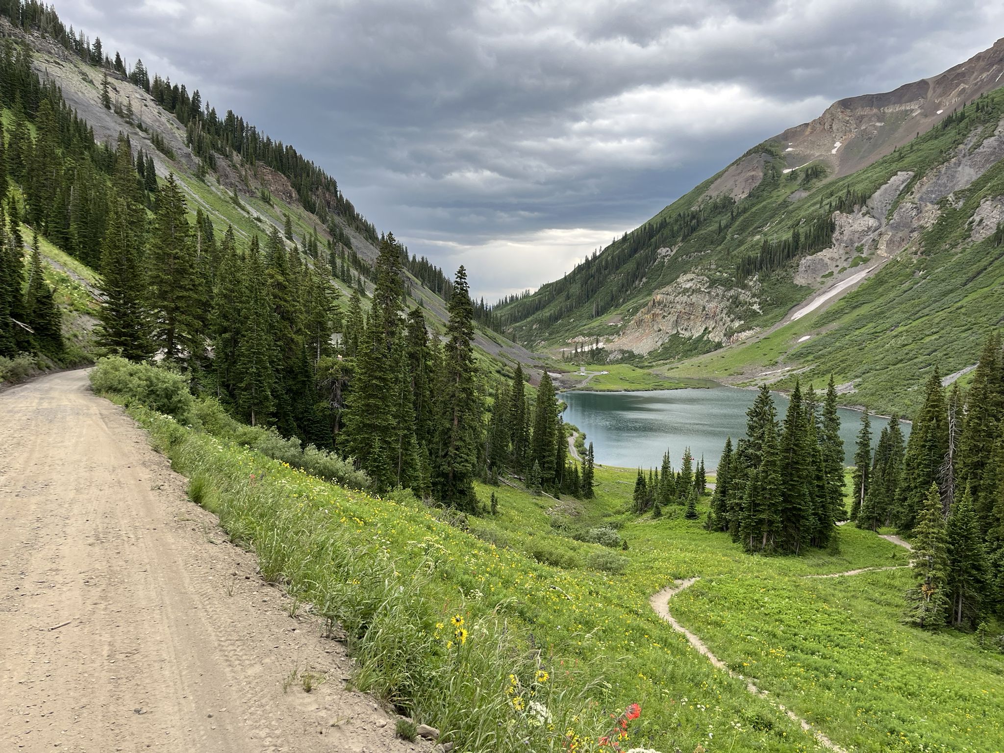 Dirt road overlooking a mountain lake surrounded by green slopes under cloudy skies. - 401 Trail Loop