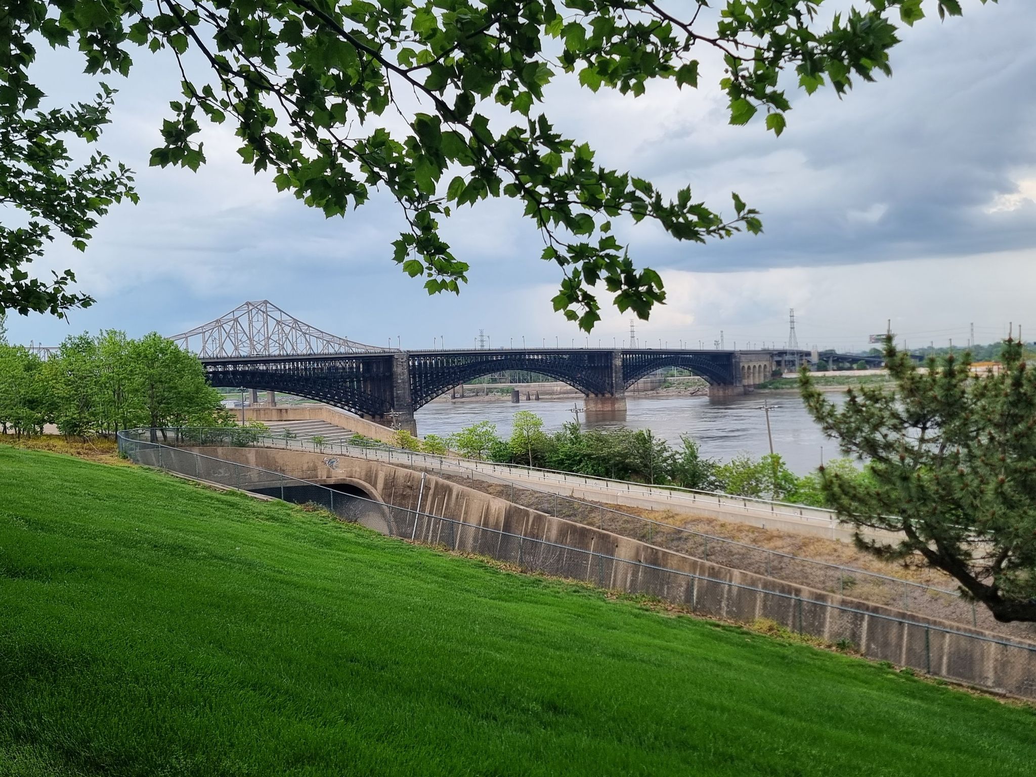 Large bridge spanning a wide river, with green slopes and cloudy sky. - Luther Ely Smith Square – Eads Bridge loop from Union Station