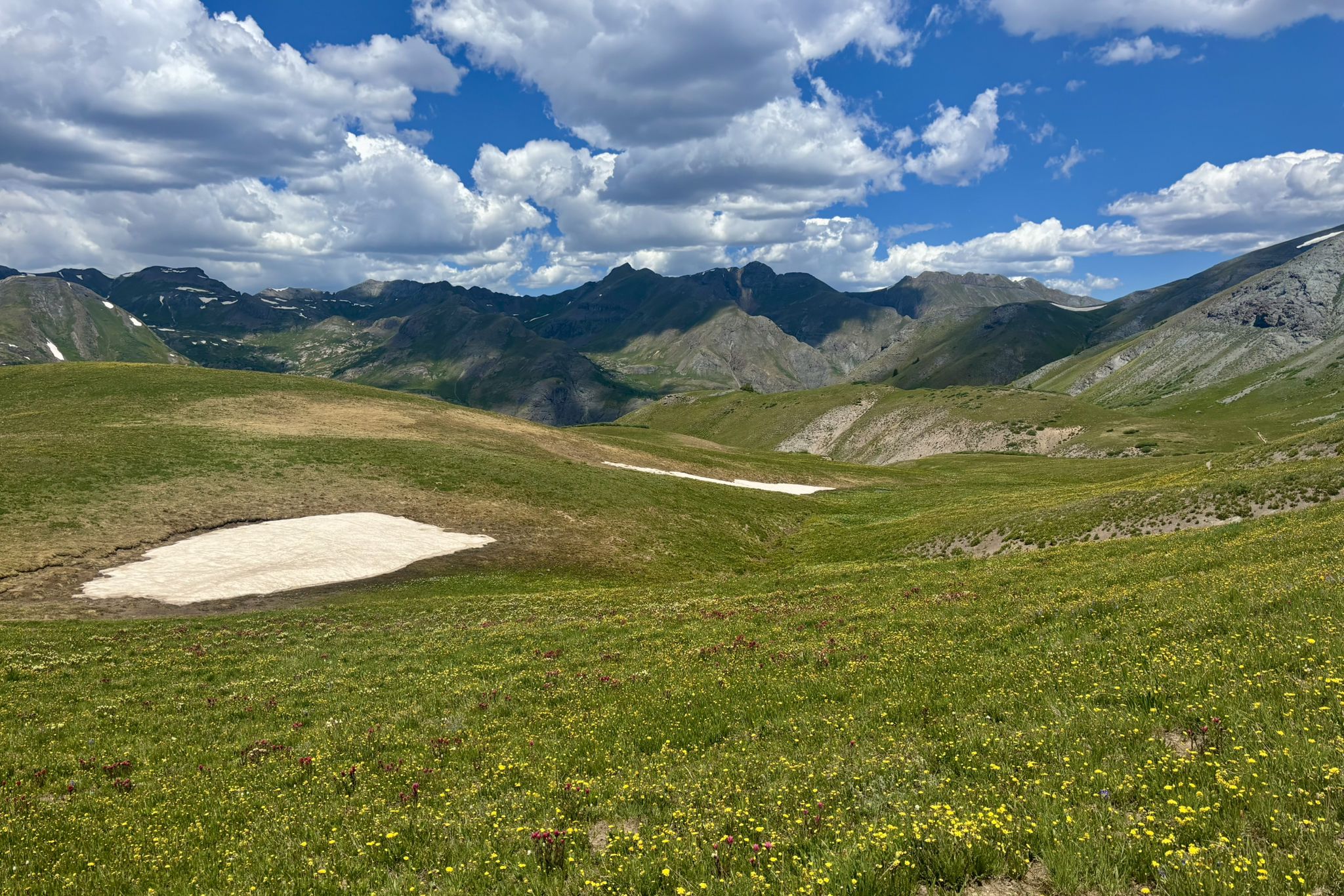 Expansive green mountain landscape with snow patches under a dramatic blue and cloudy sky. - Cunningham Gulch Trail