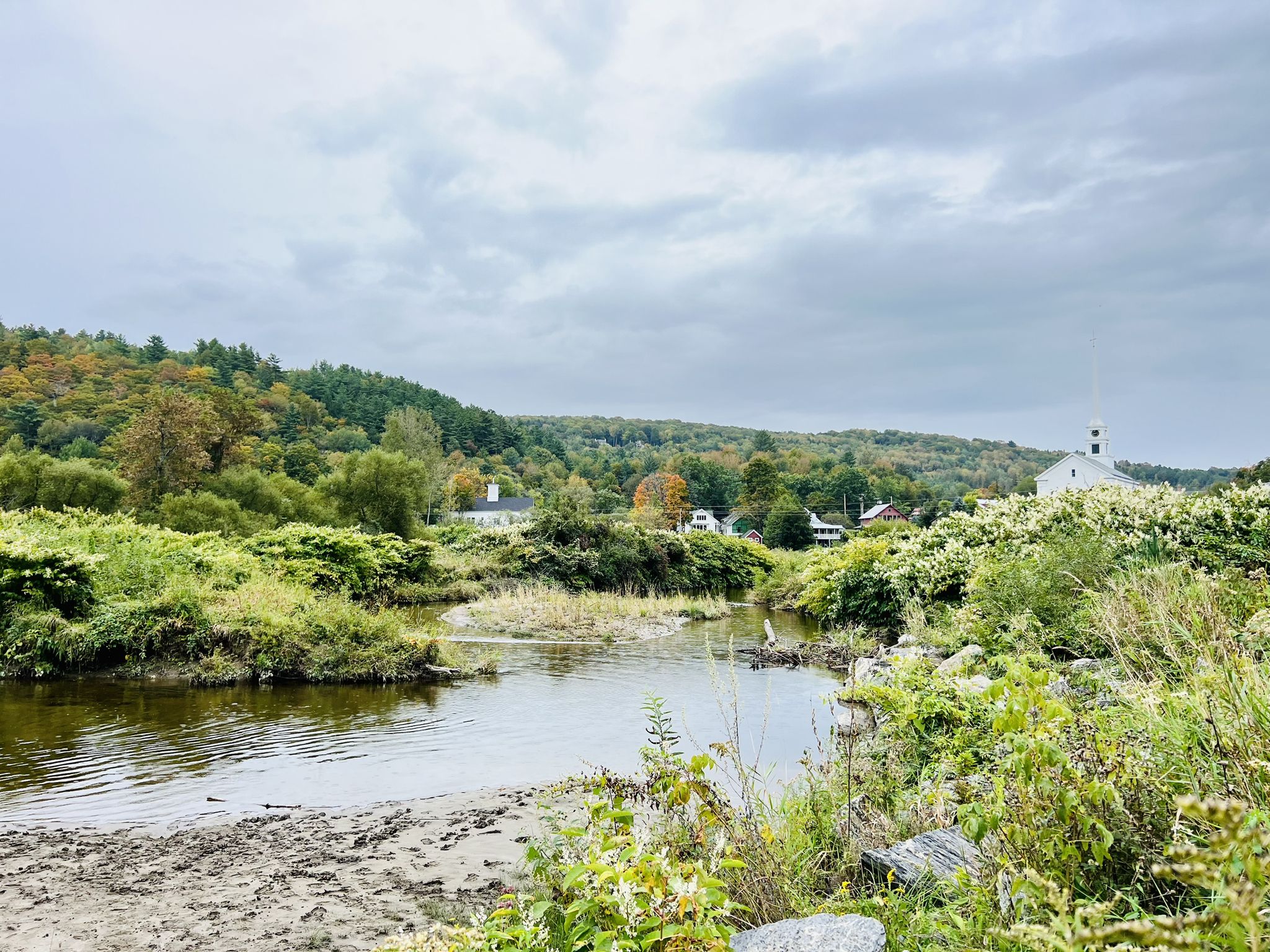 River flows through lush autumn landscape with distant houses under cloudy sky. - Gold Brook Covered Bridge – Barnes Hill loop from Trapp Family Lodge