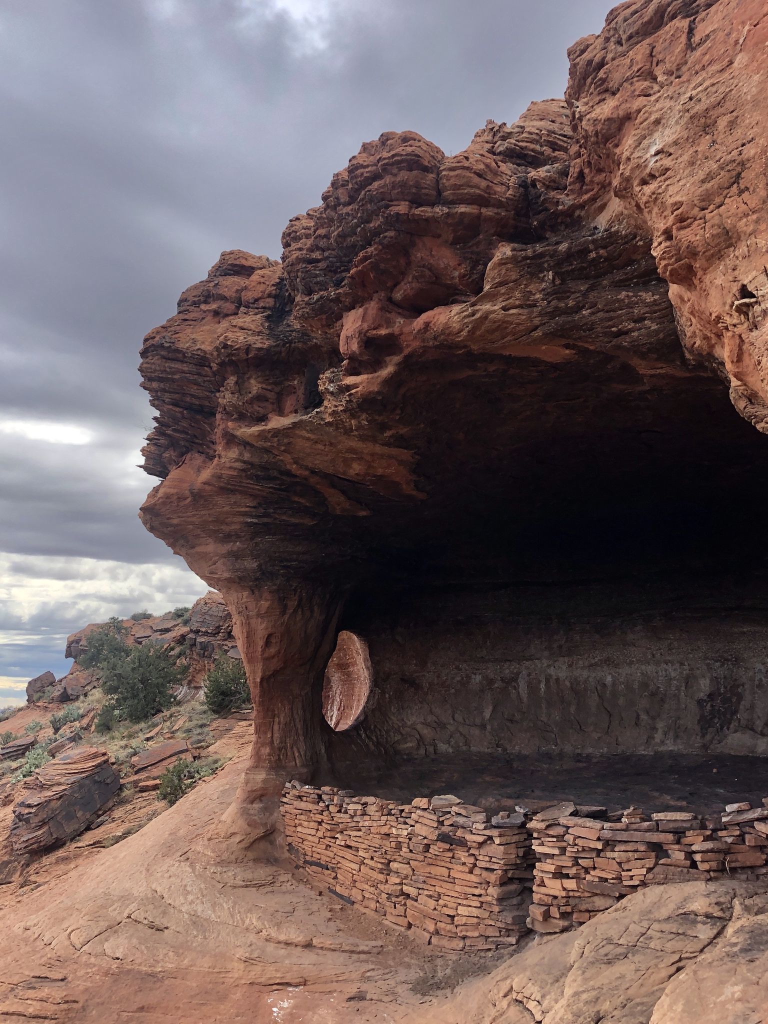 Ancient stone wall under a large red rock overhang on a cloudy day. - Robbers Roost Trail