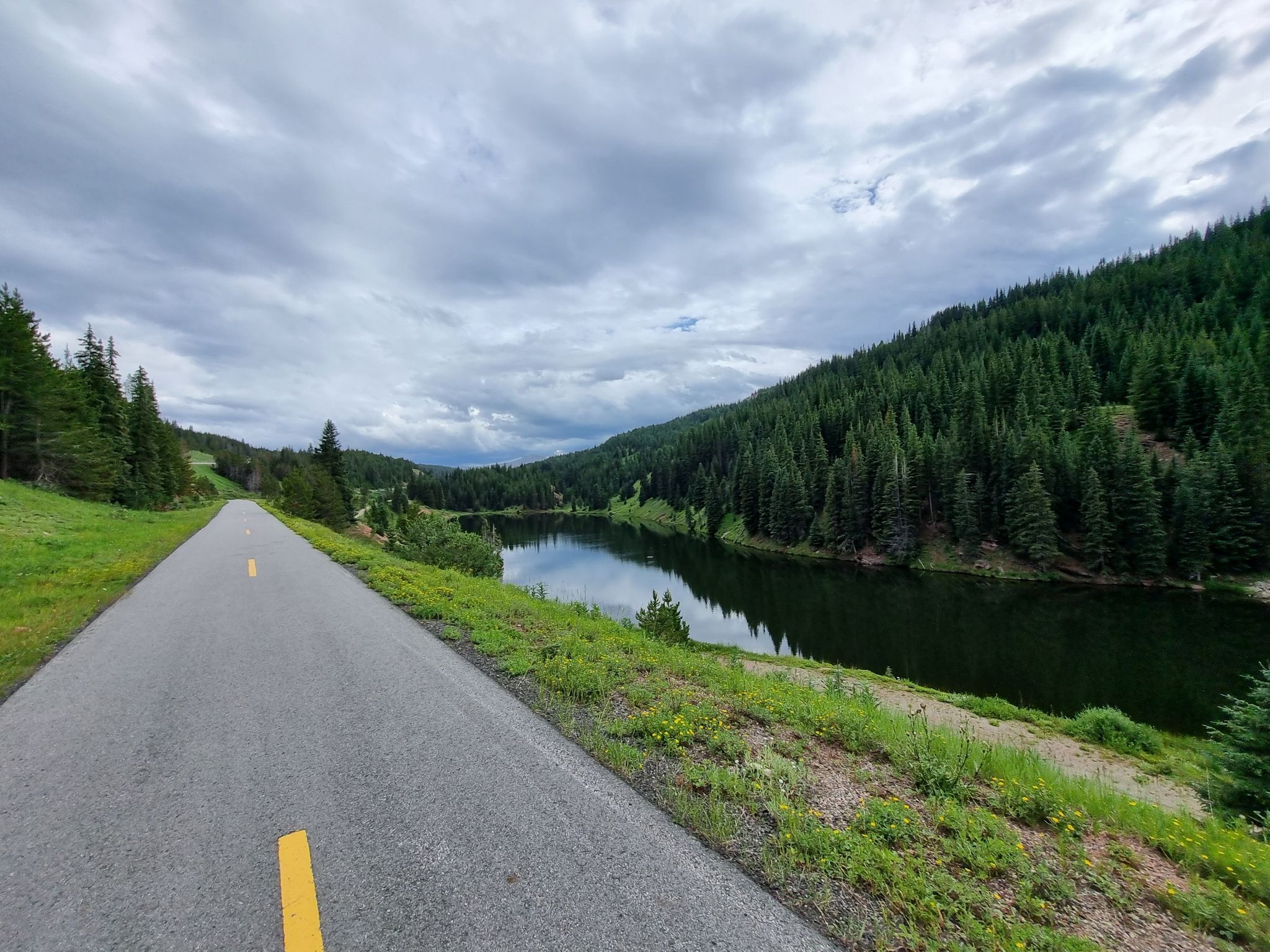 Paved road alongside a calm lake, surrounded by dense pine forests under cloudy skies. - Minturn, Red Cliff, and Vail Pass loop from Vail