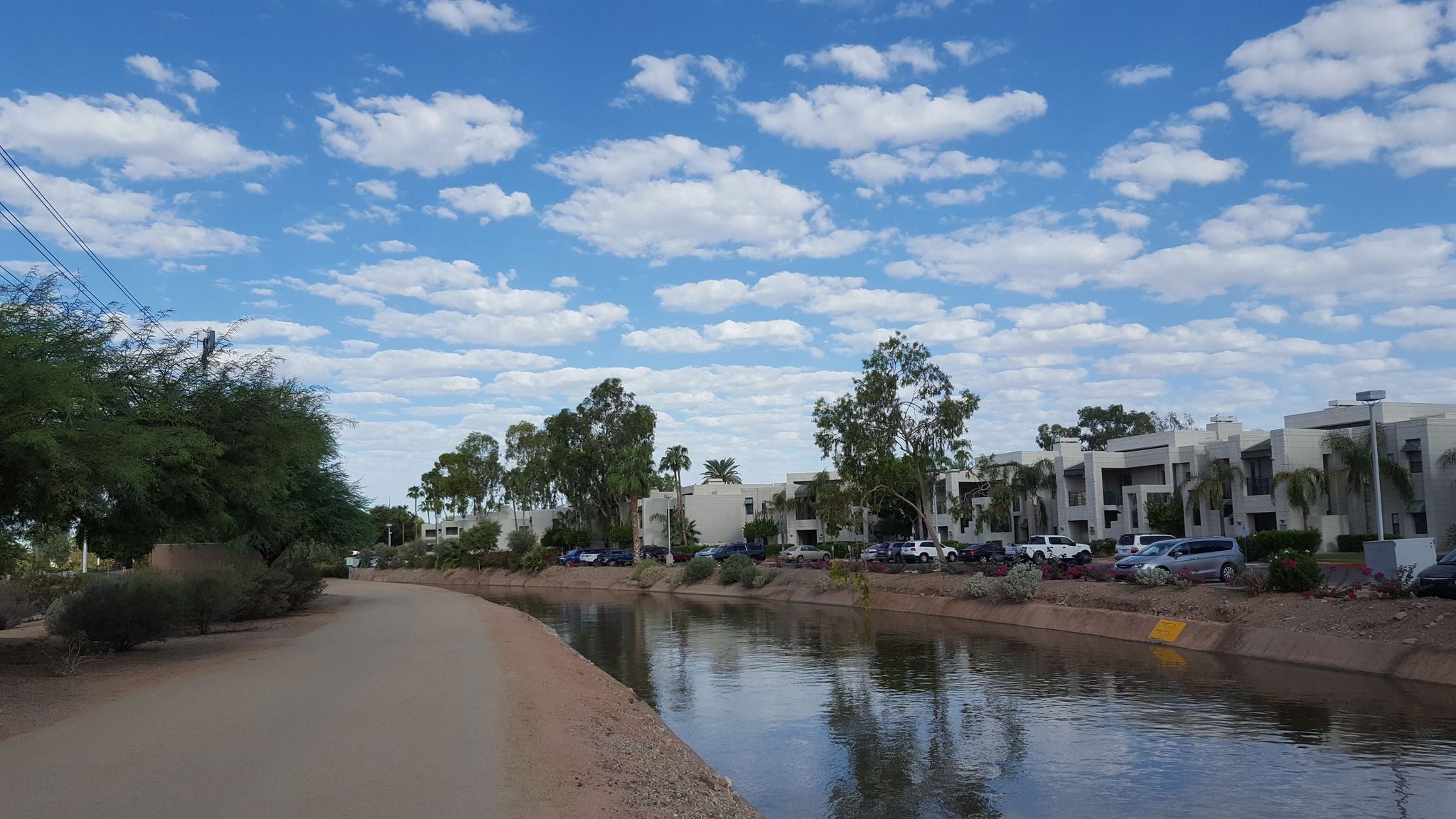 A canal with a dirt path and modern buildings under a bright, cloudy sky. - Arizona and Grand Canal Loop