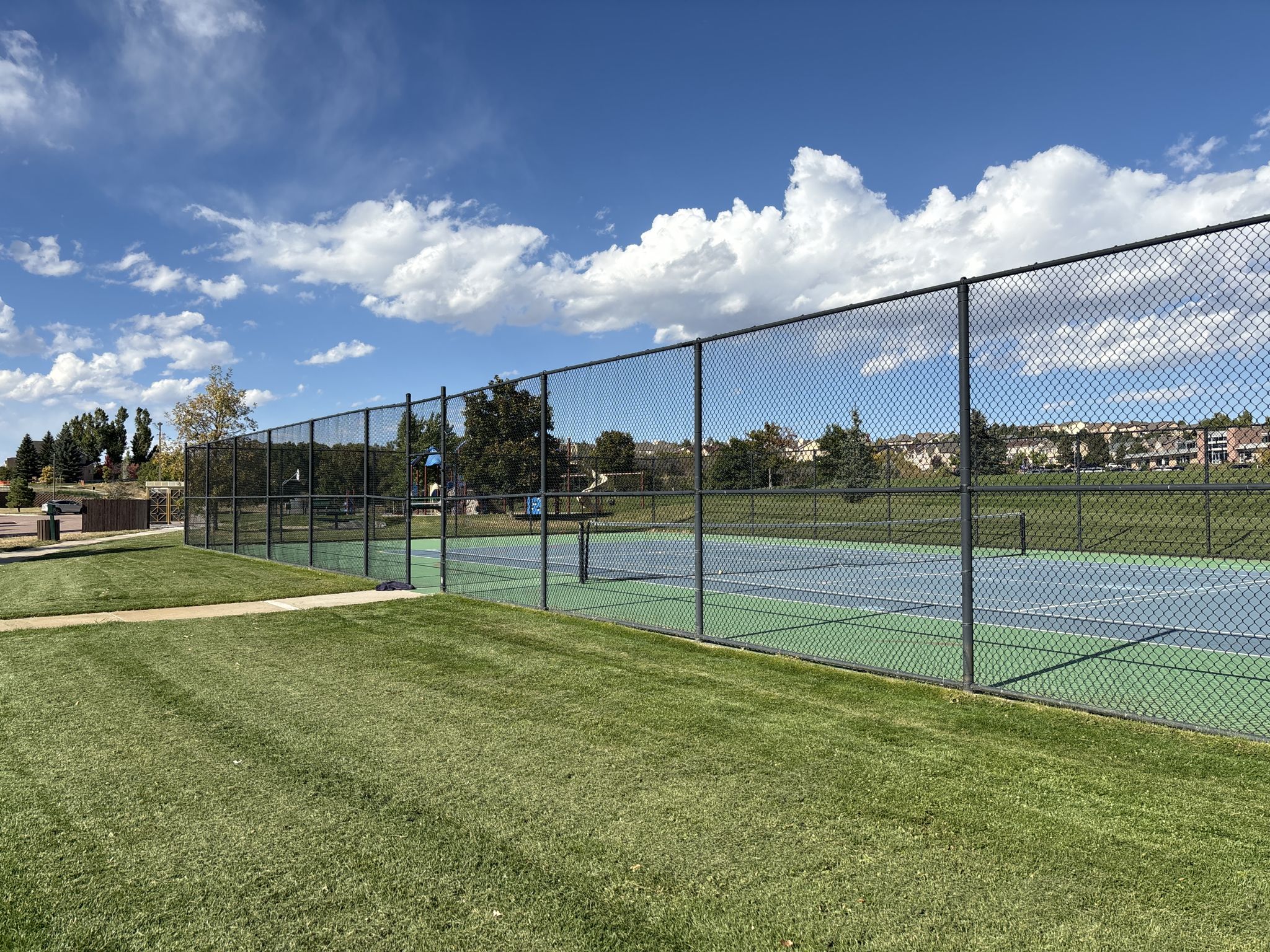 Sunny tennis courts behind a chain-link fence, with green grass and trees. - La Foret Trail