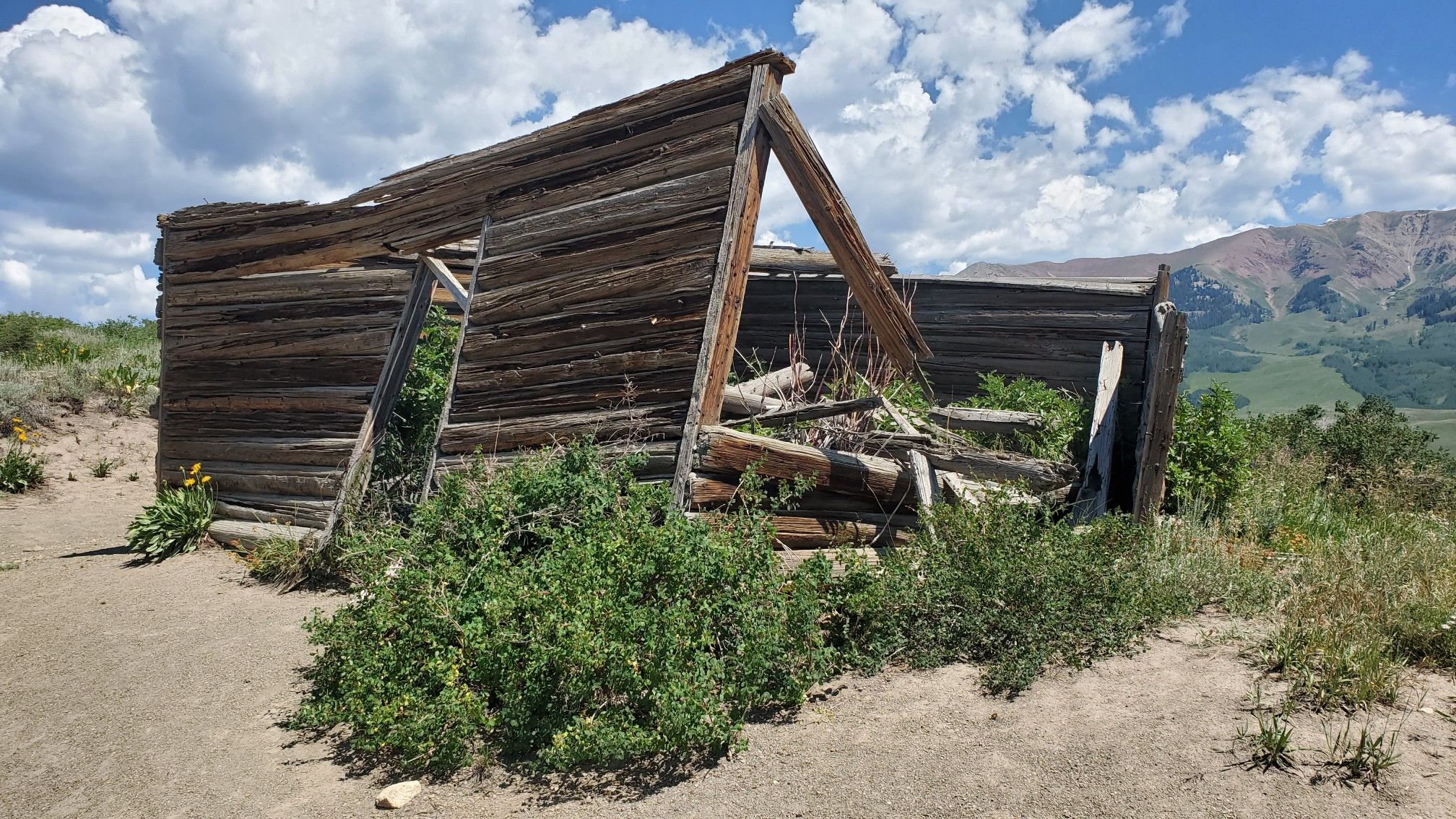 A dilapidated wooden structure stands on a grassy hill under a cloudy sky. - Teddy's Trail
