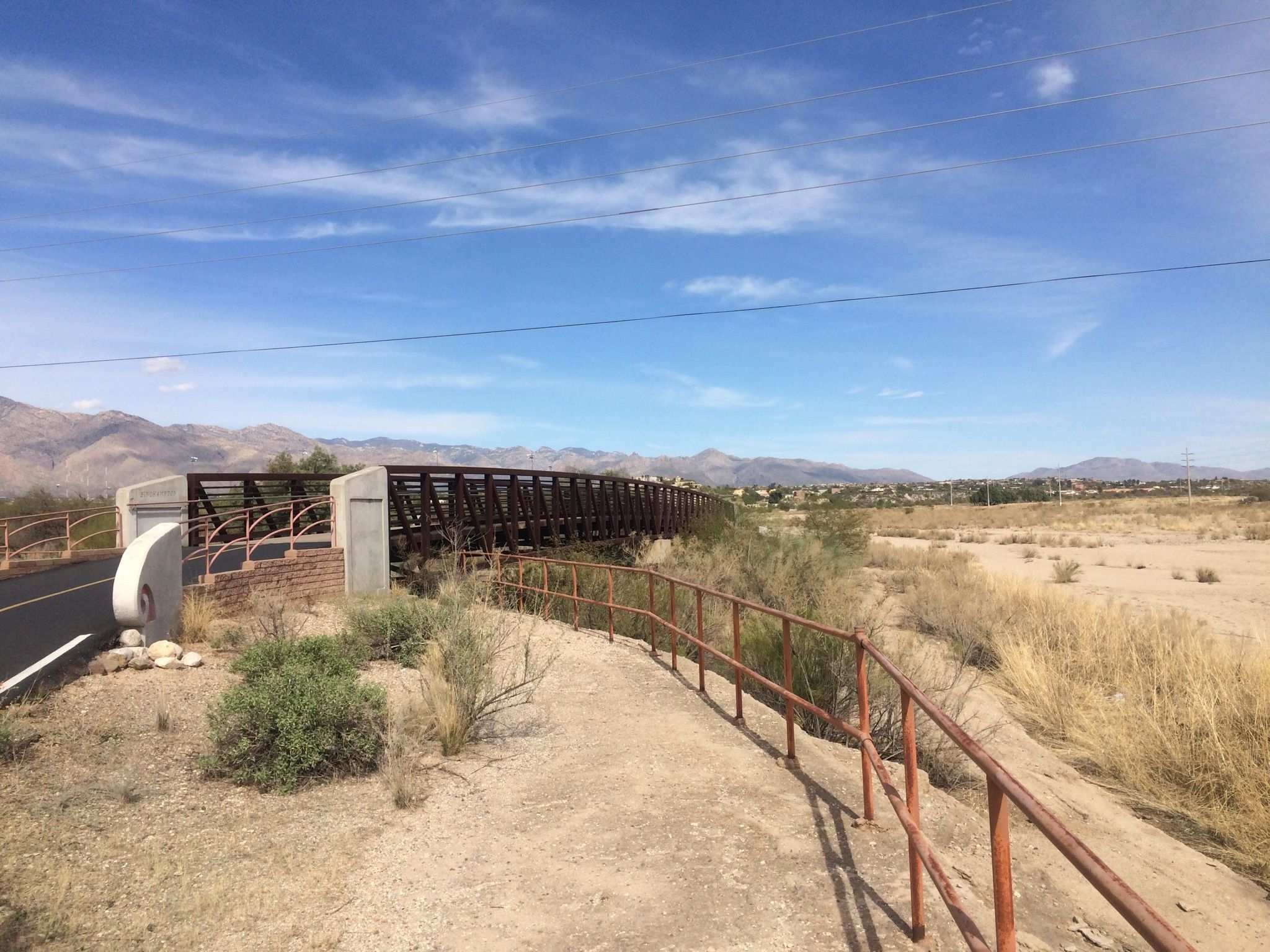 Metal pedestrian bridge over a dry riverbed, with mountains in the distance. - Wild Spirit – Rillito River Path loop from Sneed Parcel
