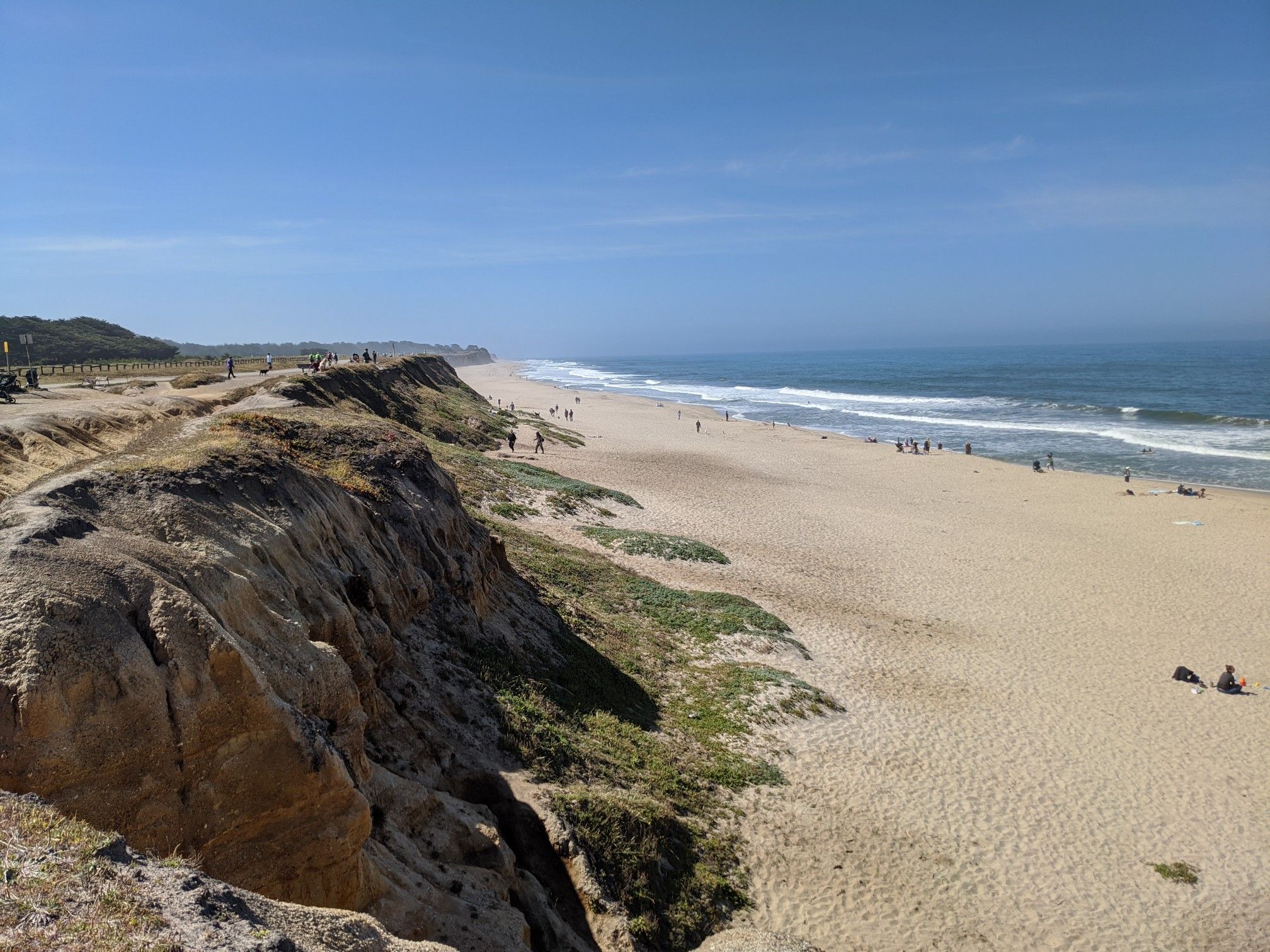 Sunny beach with sandy cliffs, ocean waves, and people enjoying the shore. - Half Moon Bay Coastal Trail