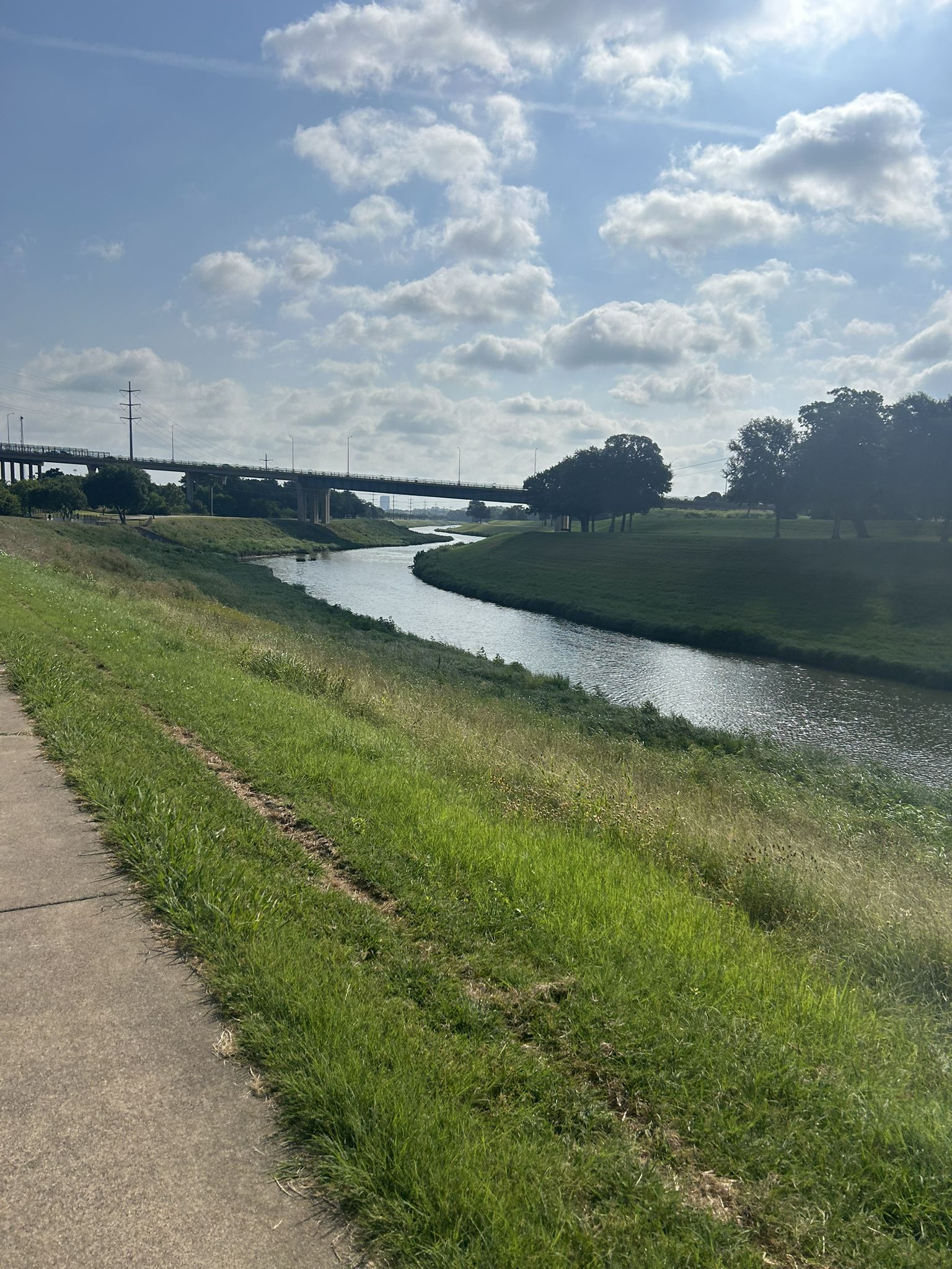 River flowing under a bridge, with a paved path and green grass. - Trinity Trails