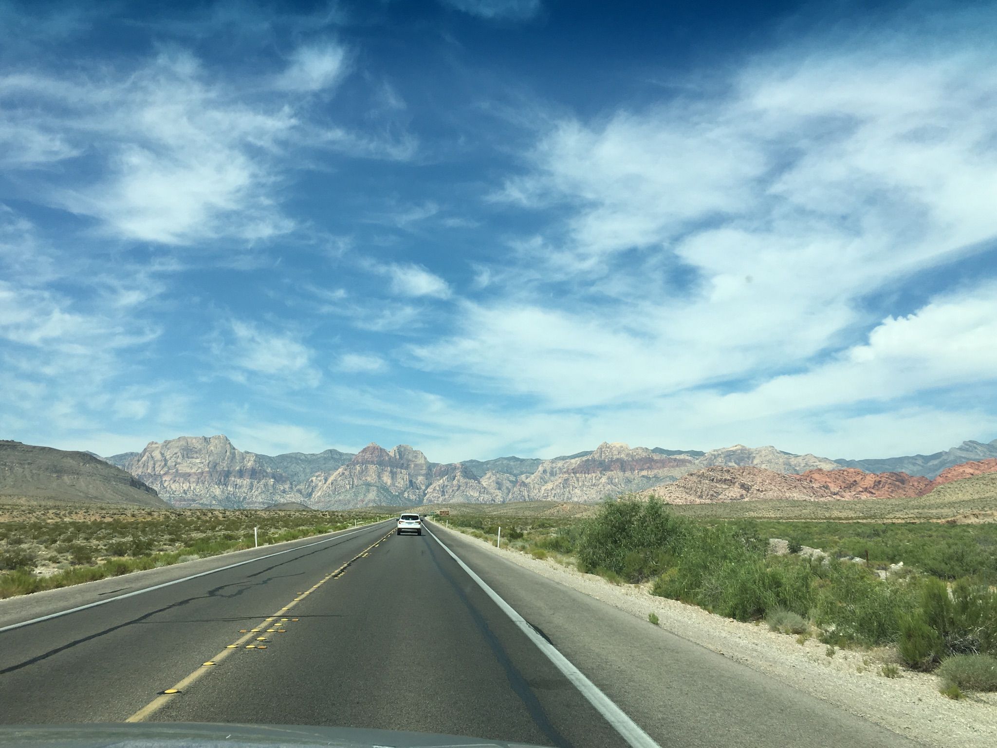 A car drives on a long desert road towards colorful mountains under a cloudy sky. - Northern Beltway Trail – Northern Beltway Trail loop from Scenic Area / Red Rock Sign