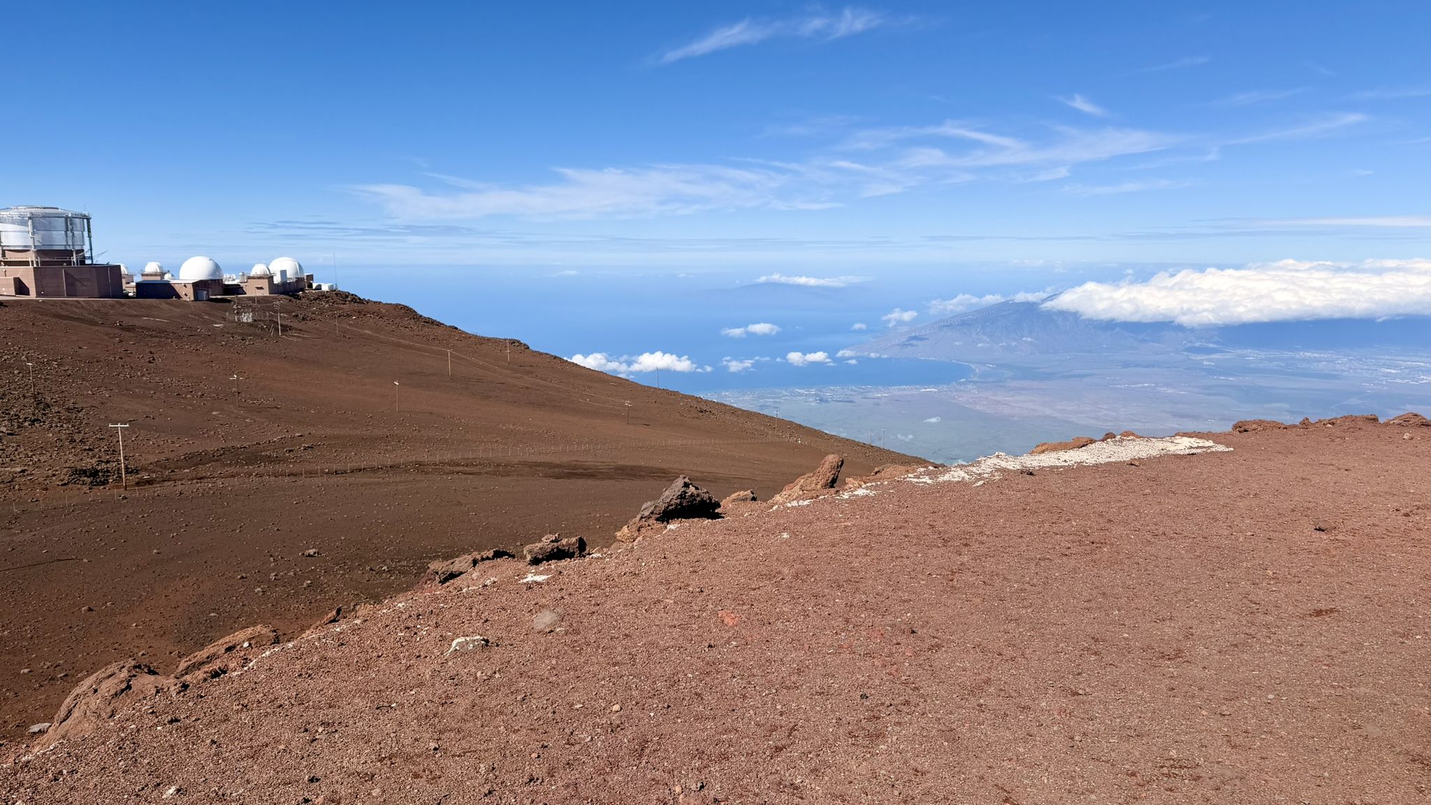 Distant observatory buildings on a brown slope overlooking clouds and distant land. - Haleakalā via the Park Headquarters Visitor Center