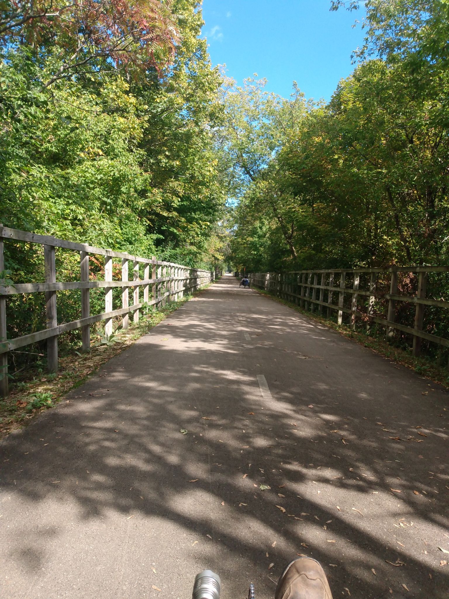 Paved path lined with wooden fences and green trees on a sunny day. - Voyageur Park – Voyageur Park loop from Greenleaf