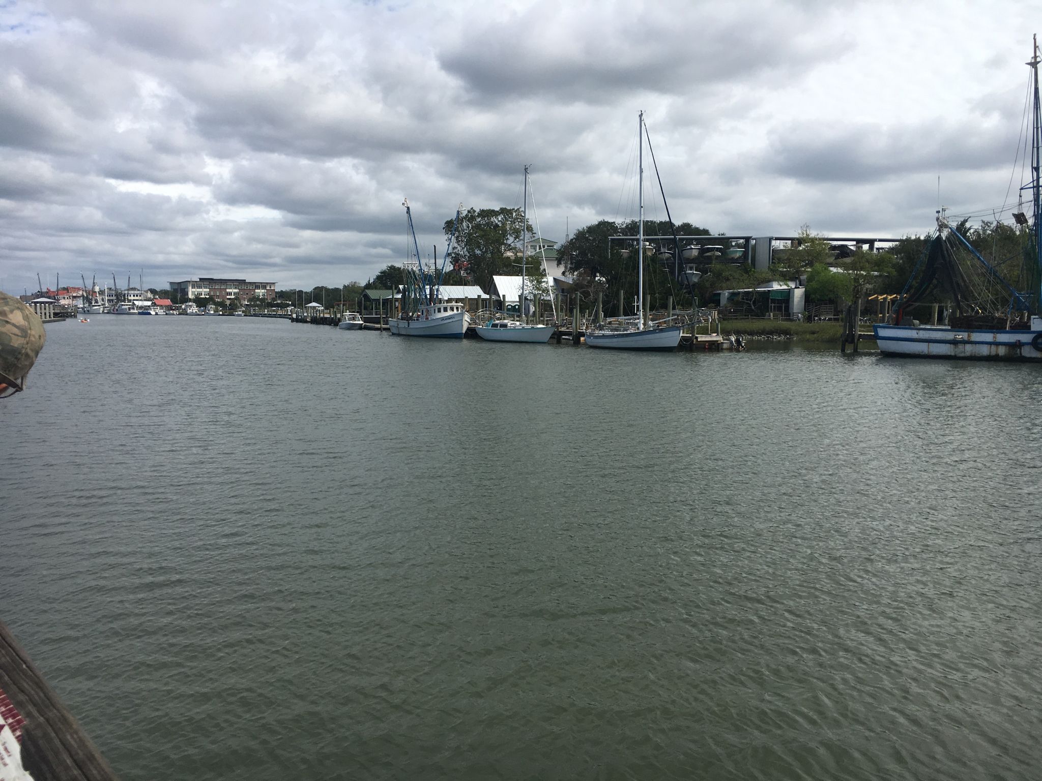 Boats docked along a wide canal with buildings and trees under cloudy skies. - Shem Creek Park – End of boardwalk loop from Mount Pleasant