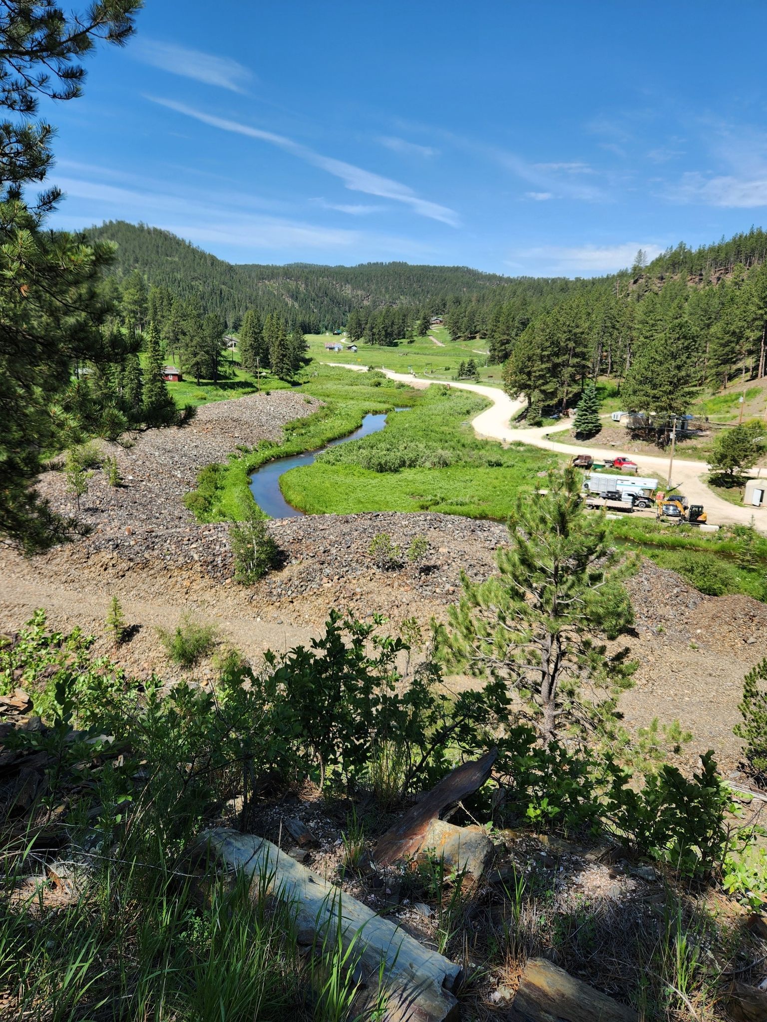 A winding river and road through a green valley with distant hills under a blue sky. - George S. Mickelson Trail