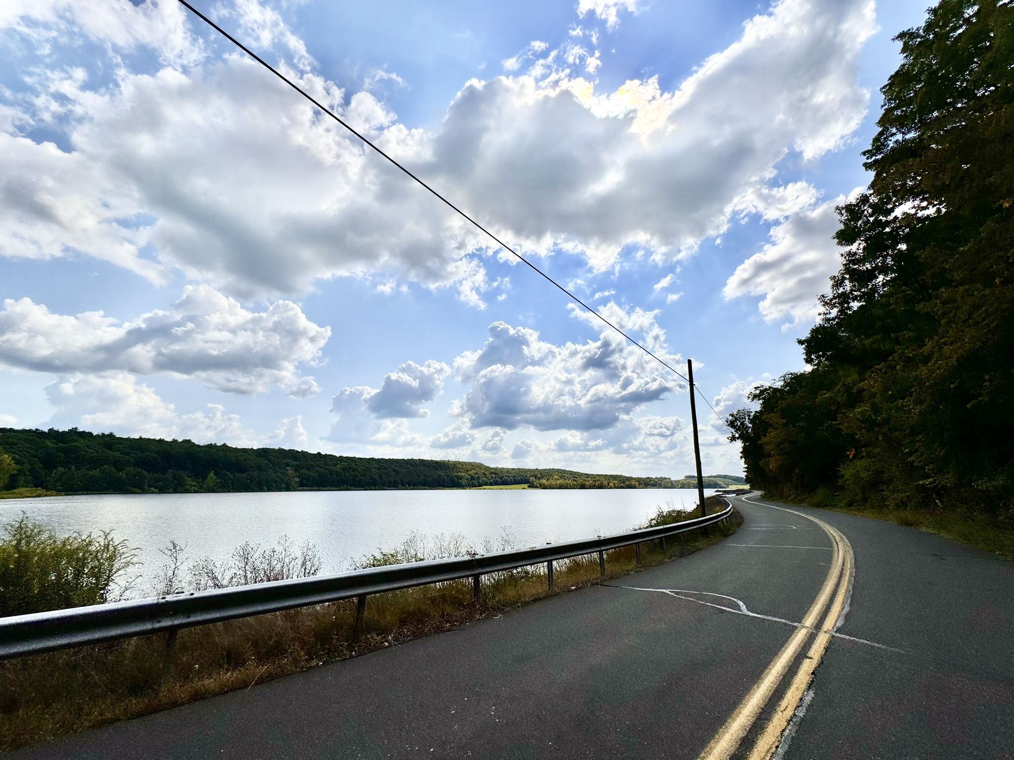 A winding road with a guardrail next to a wide body of water under a cloudy sky. - Farmington Canal Trail and Meriden Loop