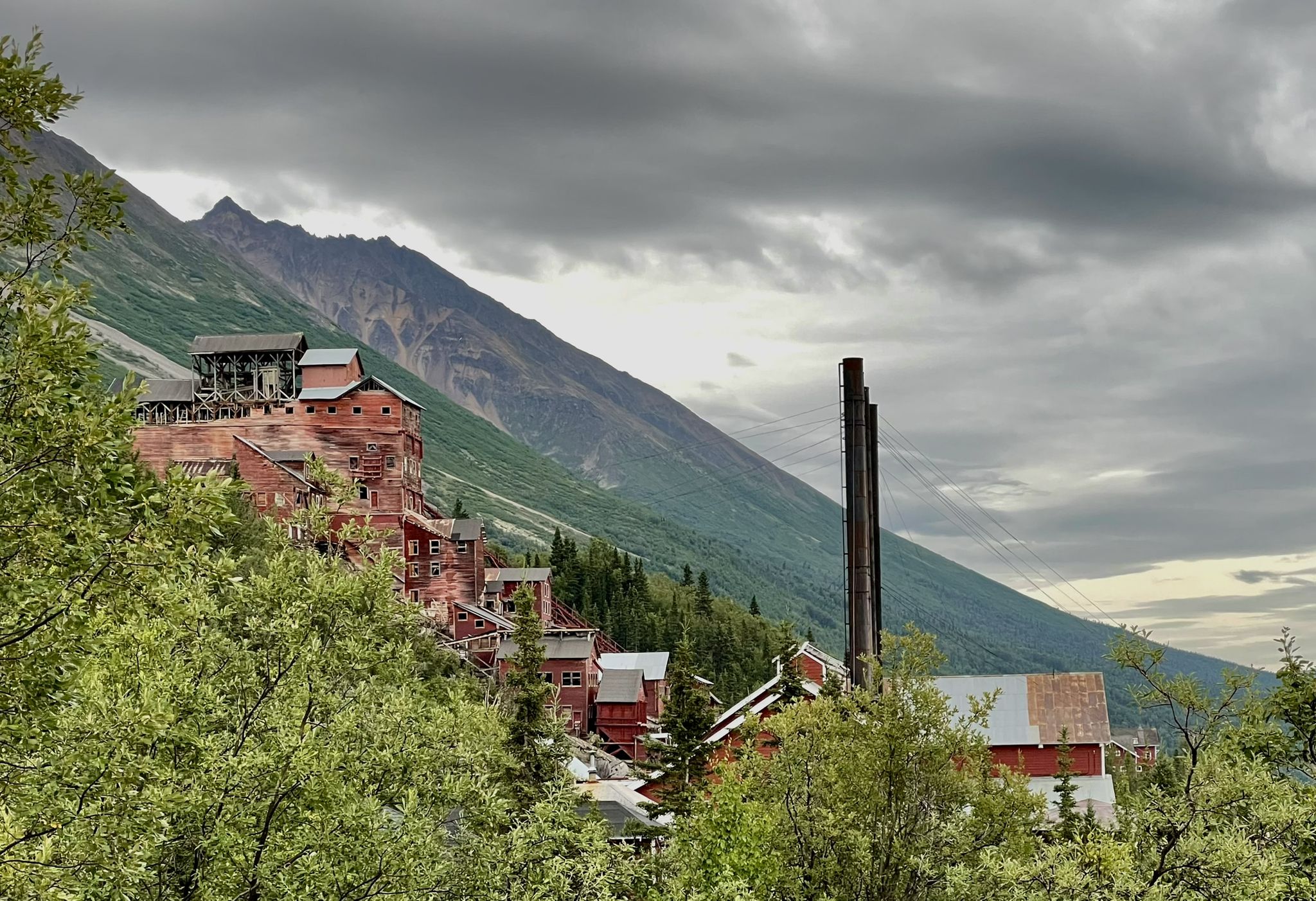 Red buildings on a mountain slope with a tall chimney under cloudy skies. - Jumbo Mine Trail