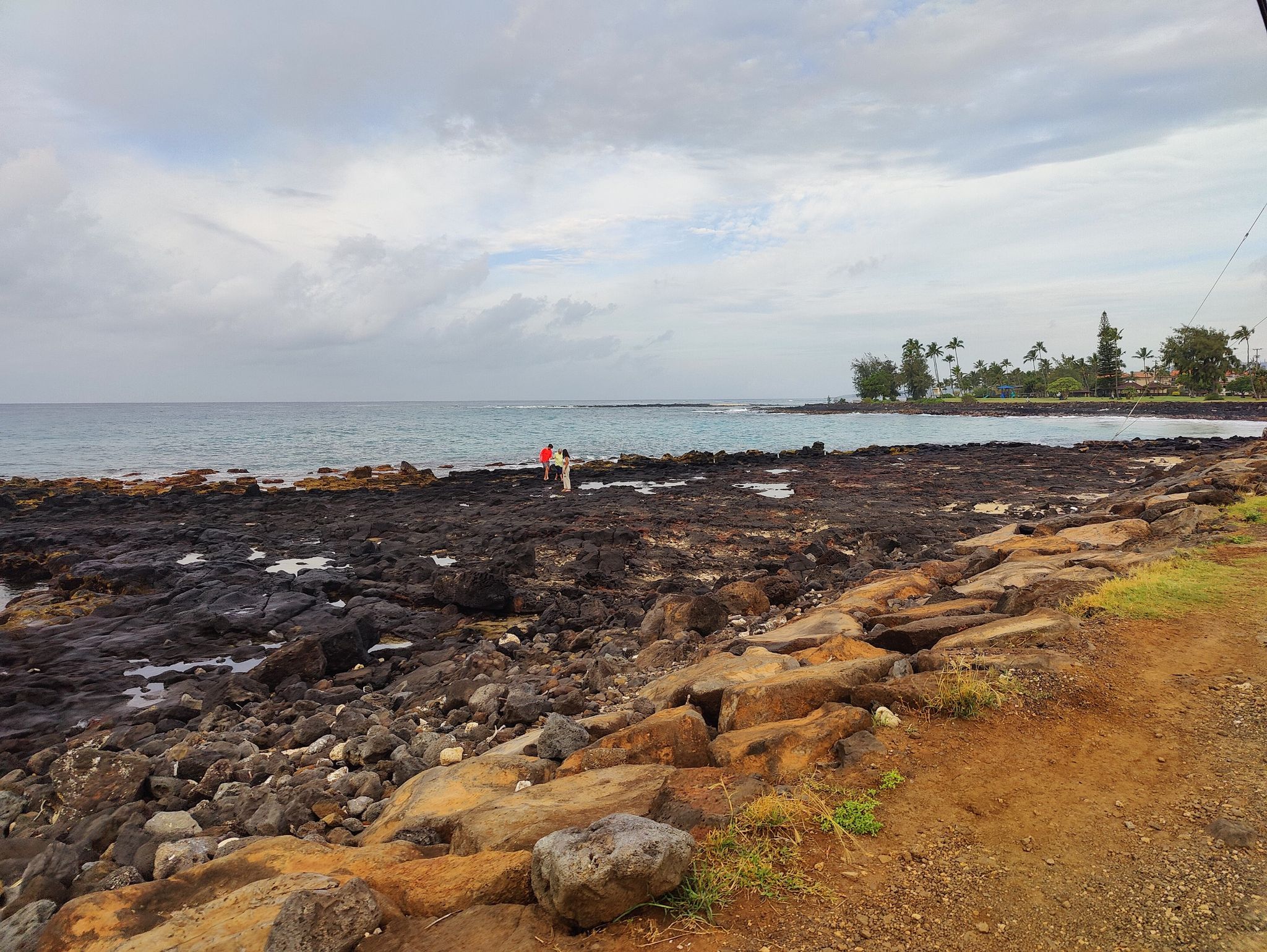 Two people on a dark rocky coastline next to the ocean under a cloudy sky. - Koloa Heritage Trail and Poipu Loop
