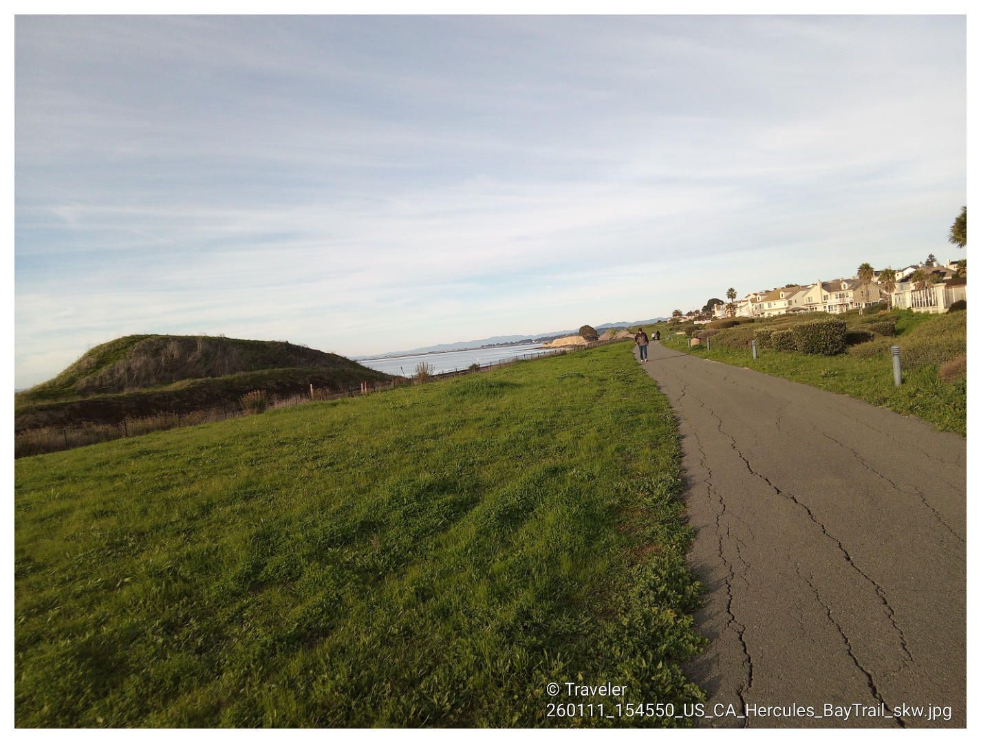 Paved path alongside a grassy hill with distant water and houses. - San Francisco Bay Trail