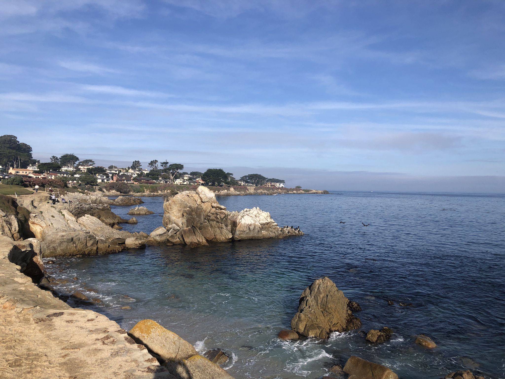 Rocky coastline with large formations, calm ocean, and distant houses under blue sky. - Cannery Row – Lovers Point Beach loop from Monterey