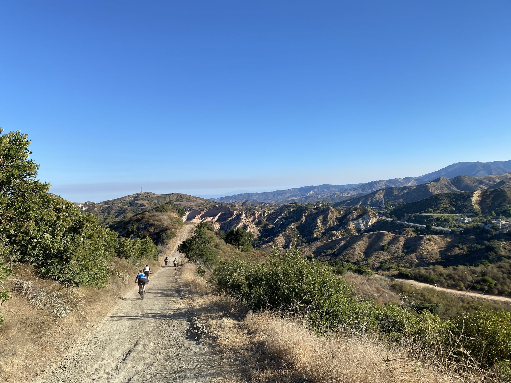 People biking on a dirt trail through dry, rolling hills under a clear sky. - Beginner Loop