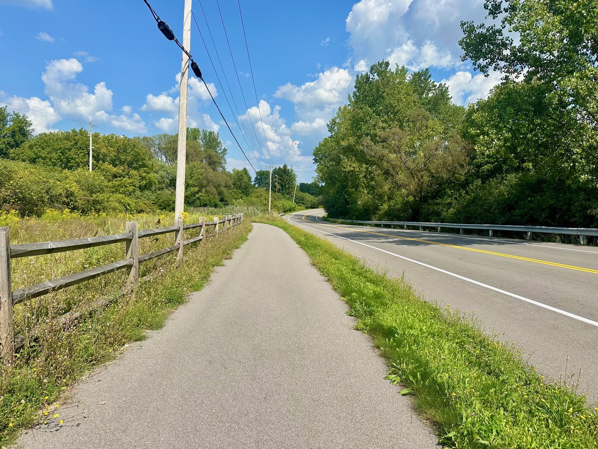 Paved path alongside a road and wooden fence, surrounded by green trees. - Erie Canalway Trail: Camillus to Syracuse