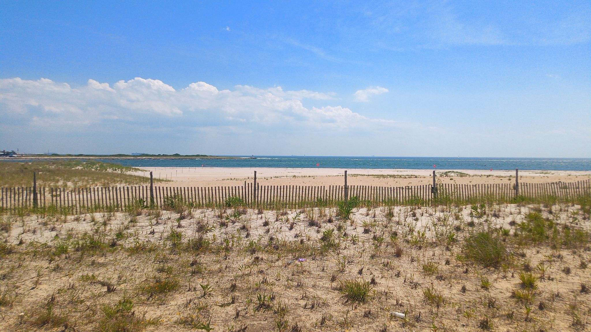 Sunny beach scene with ocean, sand dunes, and a wooden fence. - Rockaway Beach Boardwalk