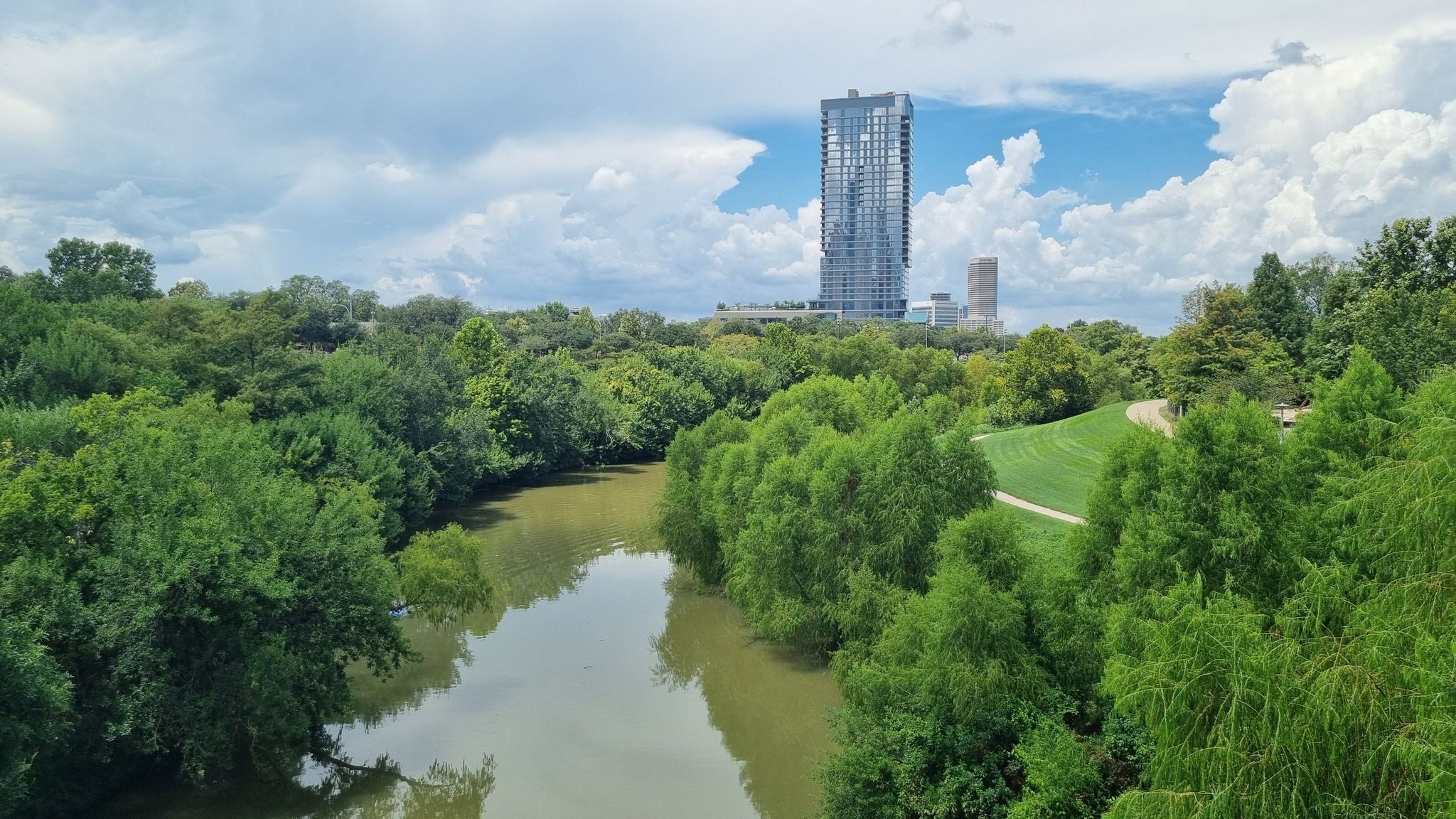 Winding river through lush green trees with a tall building under cloudy sky. - Buffalo Bayou Loop Trail
