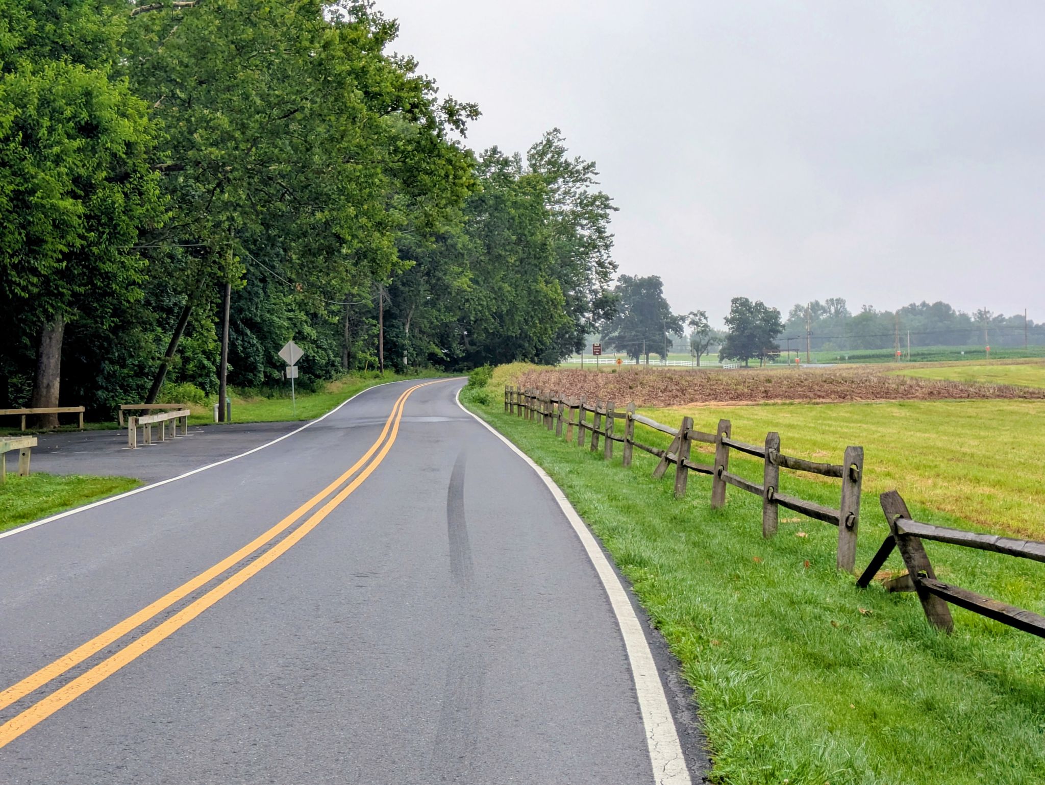 Paved road next to a wooden fence and green fields on an overcast day. - Loys Station Covered Bridge – Utica Covered Bridge loop from Emmitsburg